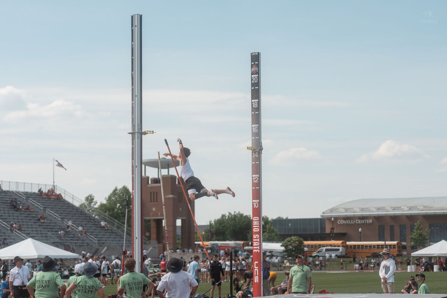Athlete performs a pole vault jump at an outdoor track and field meet with stadium seating in background.