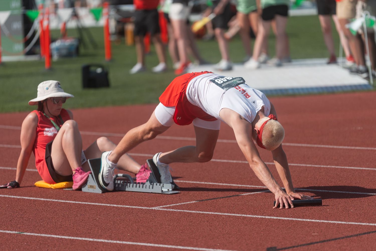 Athlete in white and red uniform sets up in starting blocks beside seated official at outdoor track meet.
