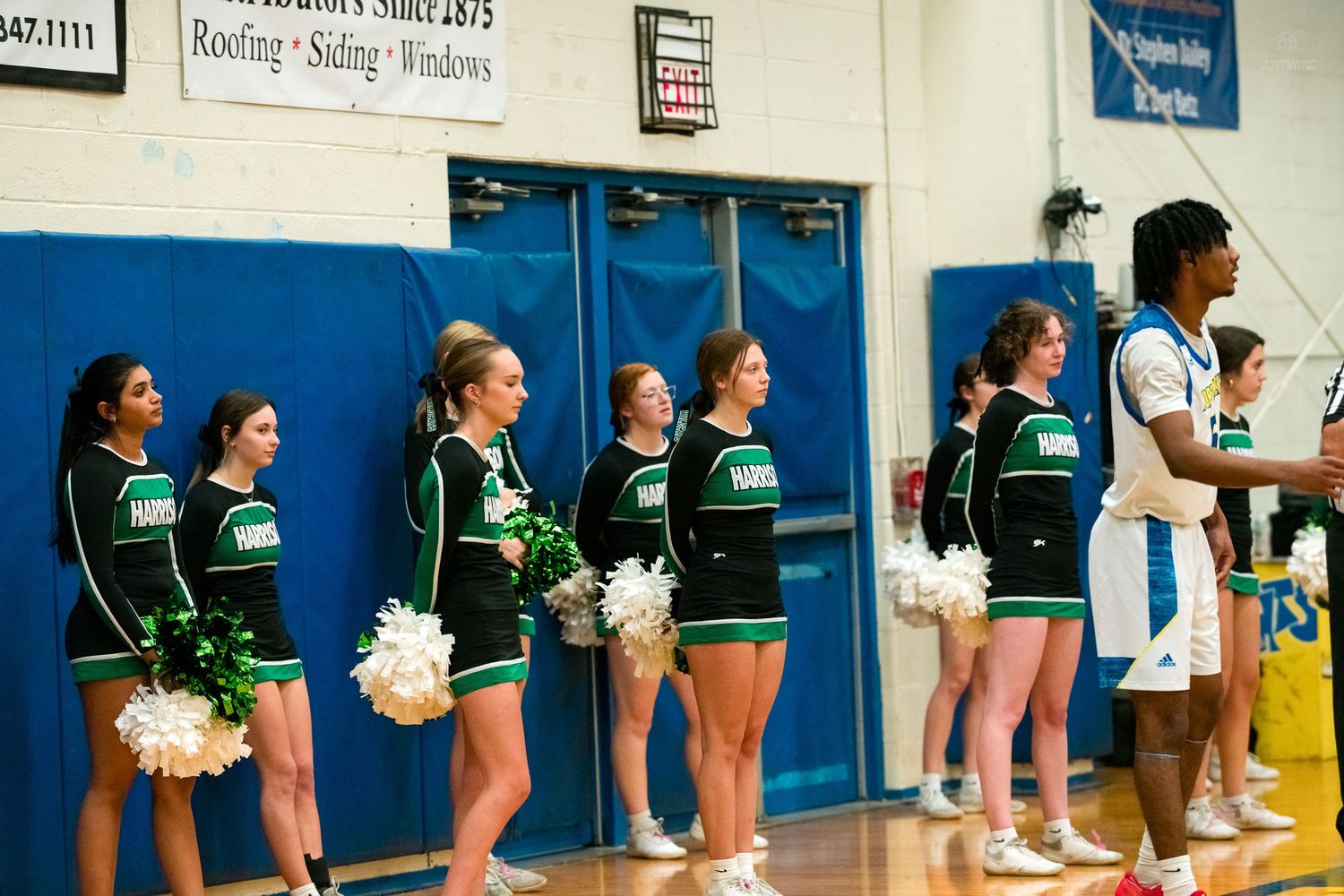 Green and white uniformed cheerleaders with pom-poms line gym wall during basketball game.