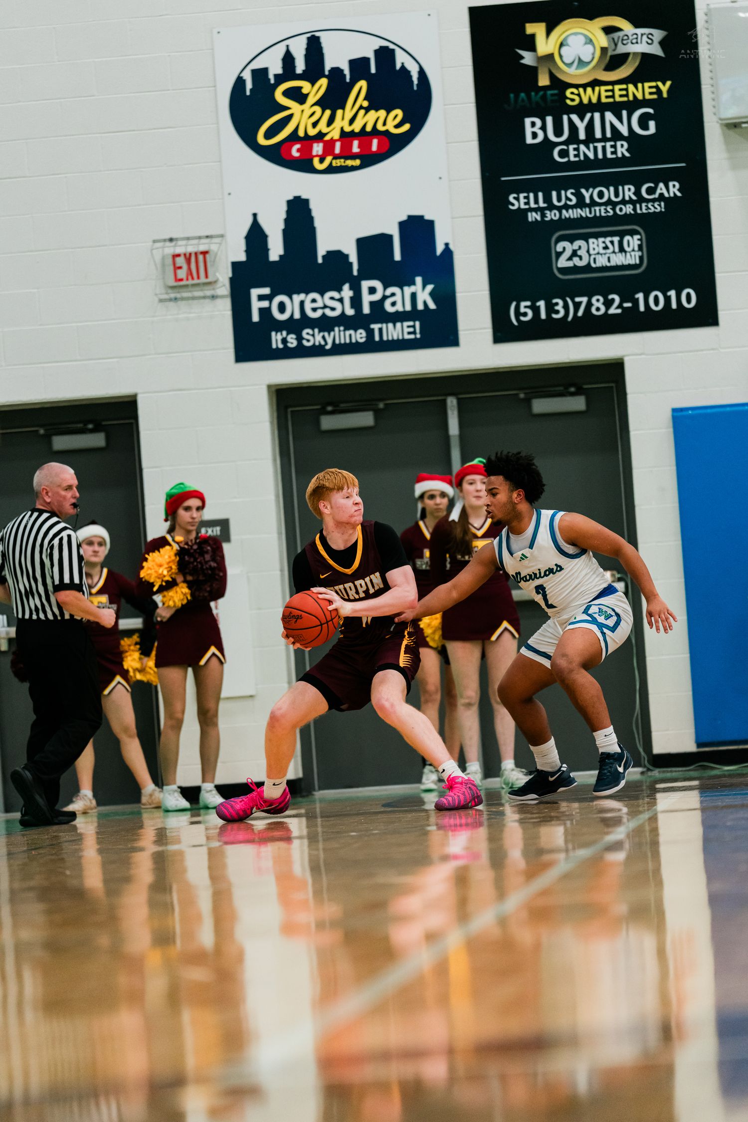 A Turpin player in maroon uniform protects the ball while being guarded by a defender in a blue home gym.