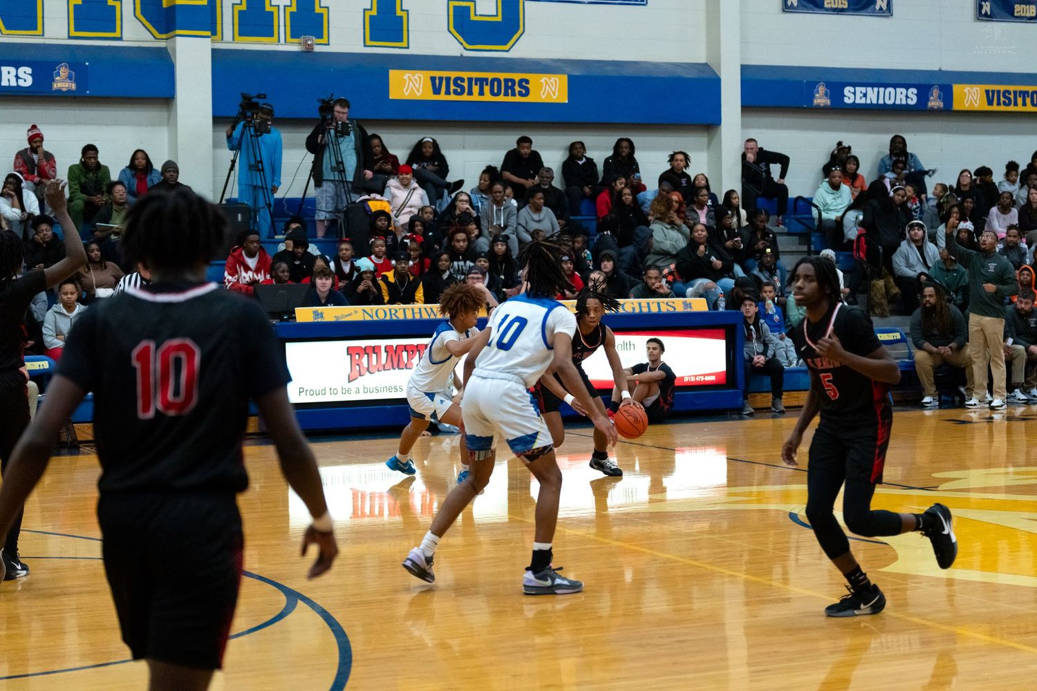 Player number 10 in blue dribbles past defenders during a high school basketball game in a packed gymnasium.