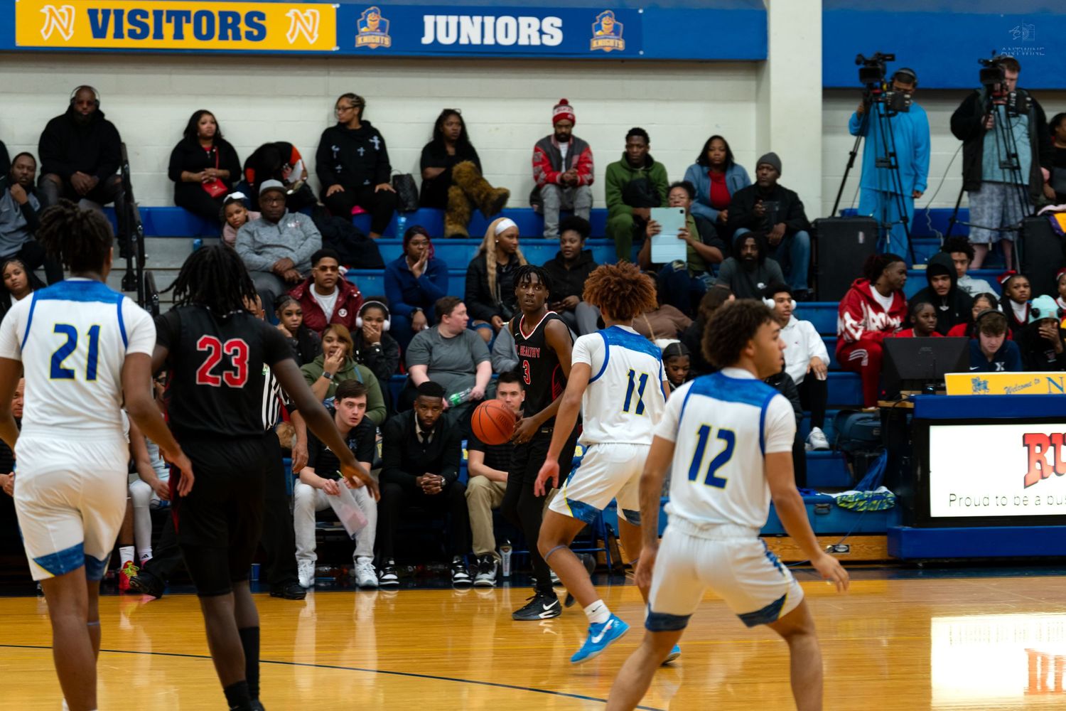 High school basketball players compete on court with crowded bleachers visible in background.