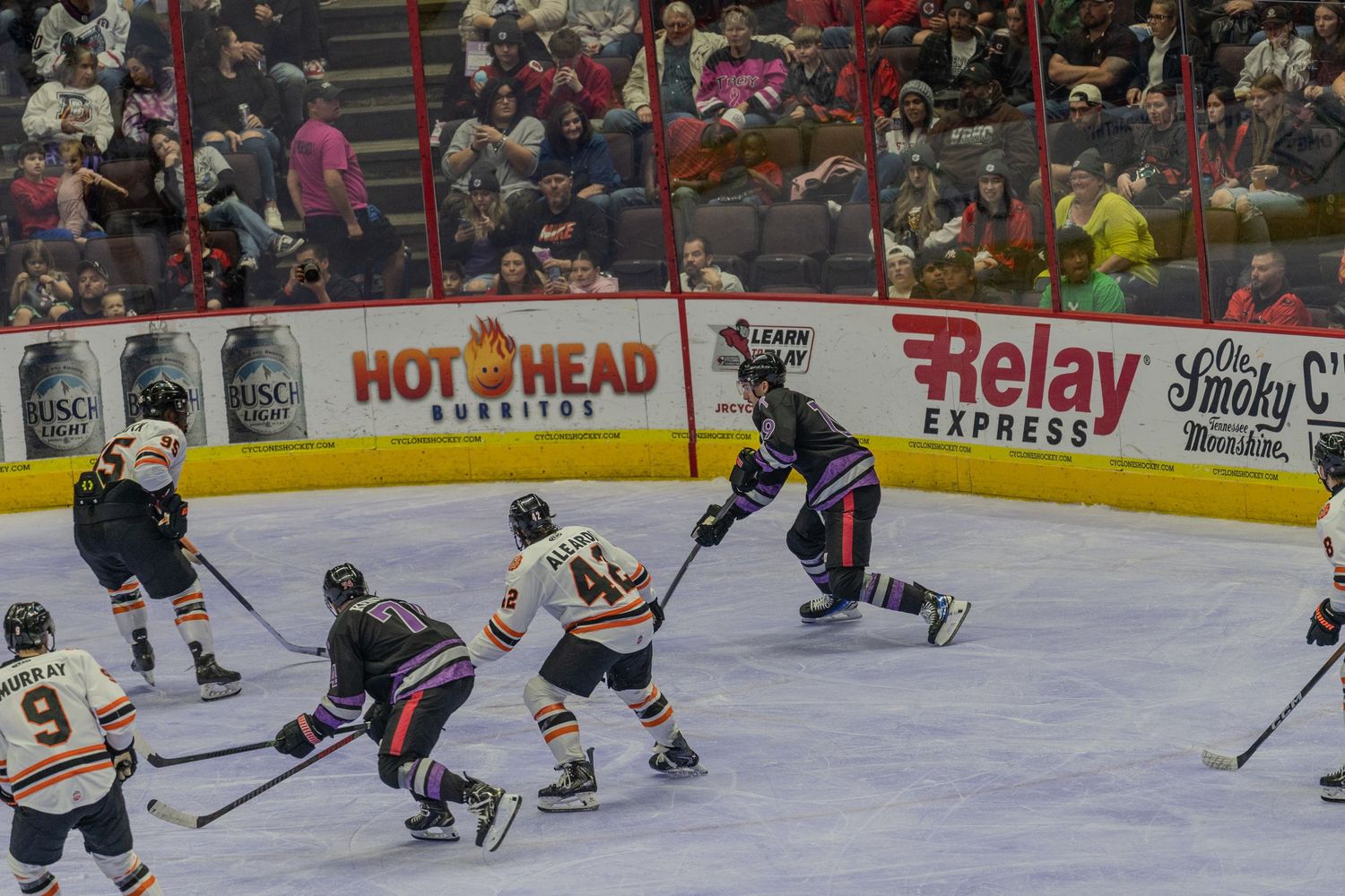 Ice hockey players in orange and purple uniforms compete for puck possession during an indoor arena game.