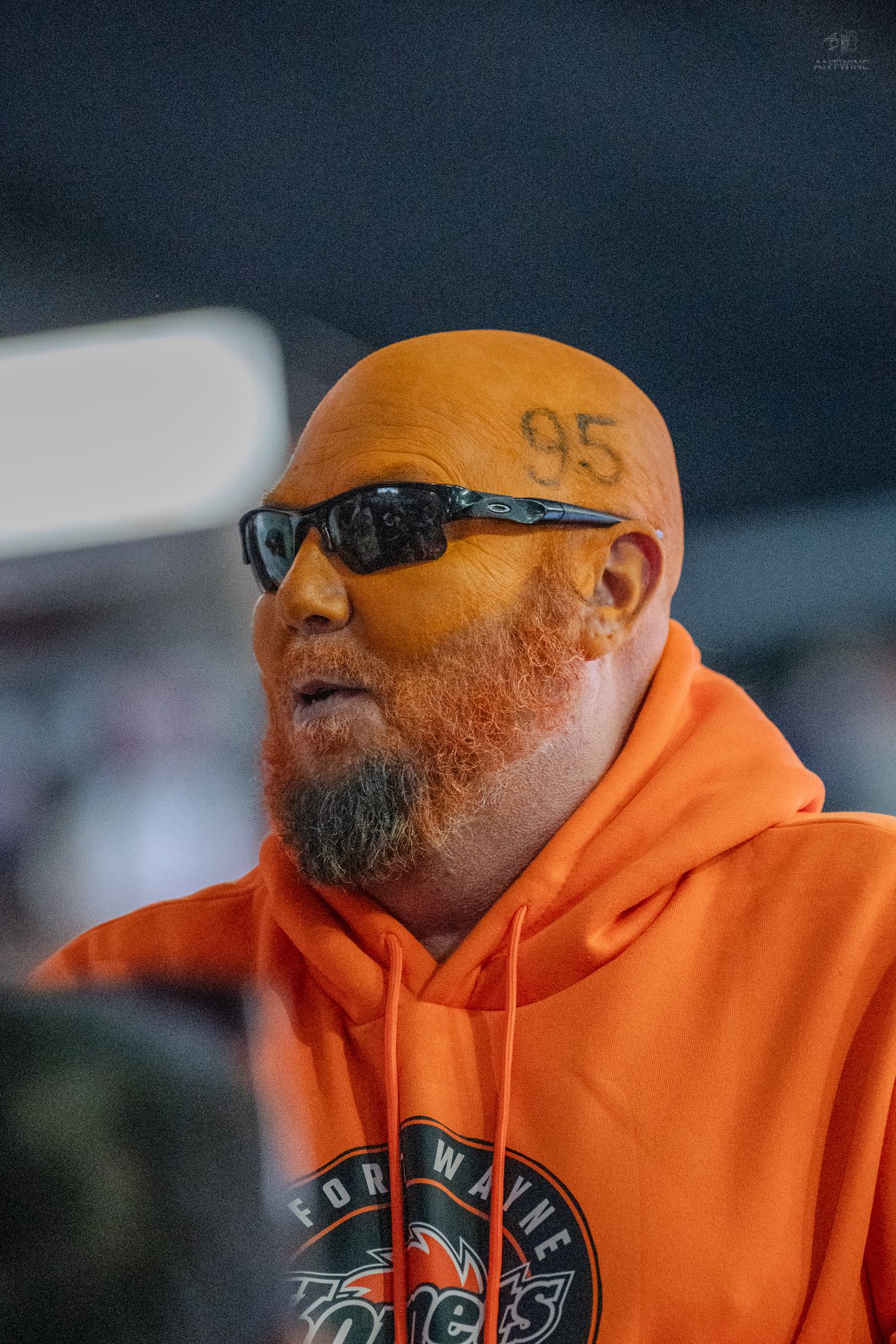 A bald man with '95' tattooed on his head wearing an orange Fort Wayne Komets hoodie and sunglasses at a hockey game.