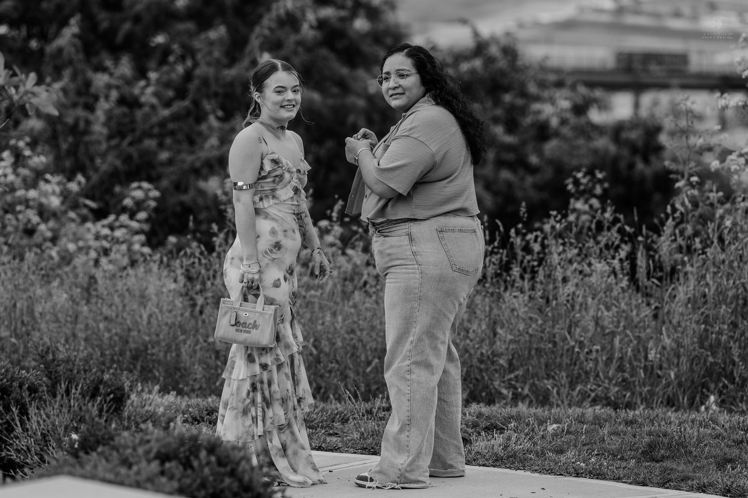 Two smiling women pose together on a garden path in this black and white outdoor portrait photo.