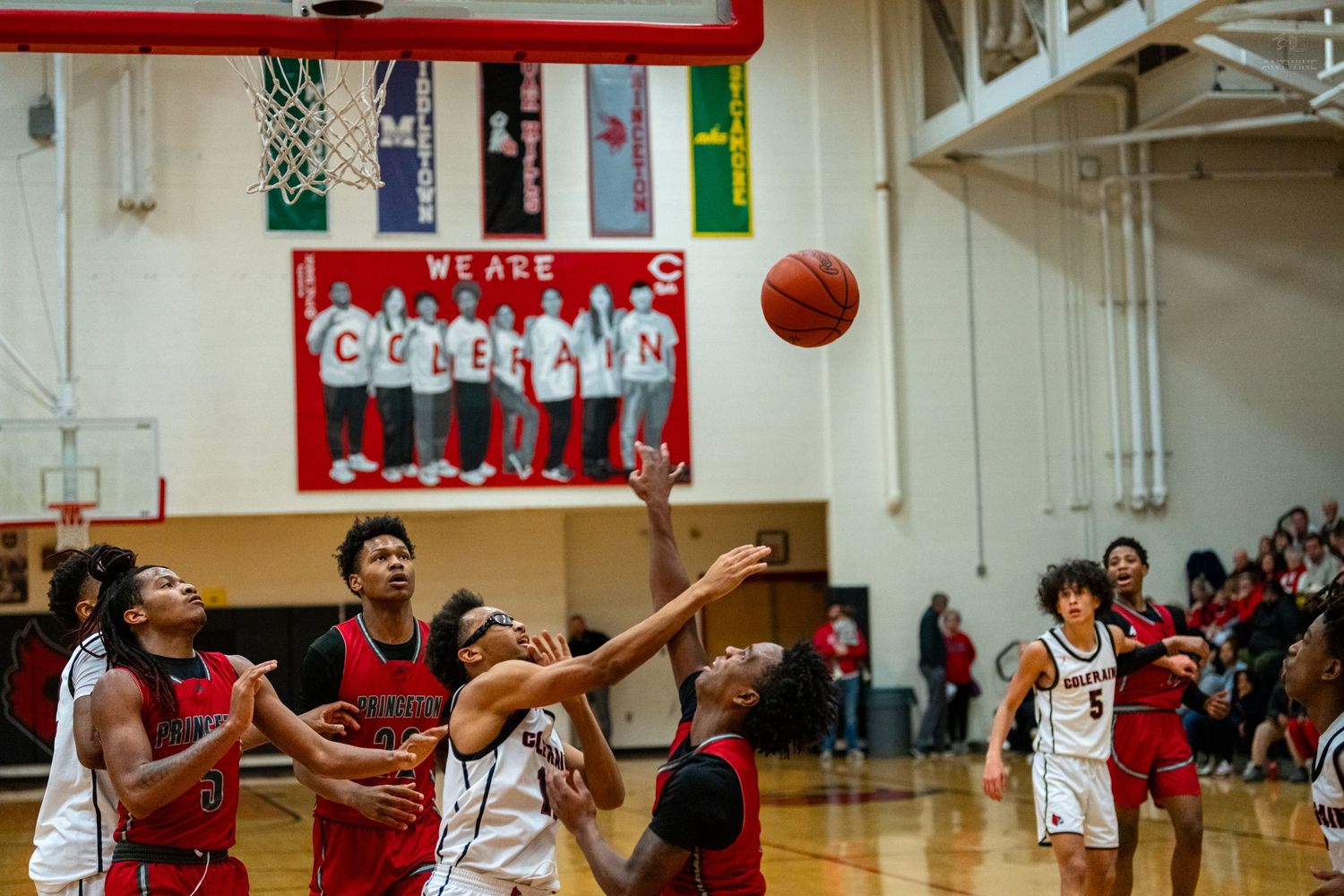 High school basketball players in red and white uniforms jump for rebound under the net during game.