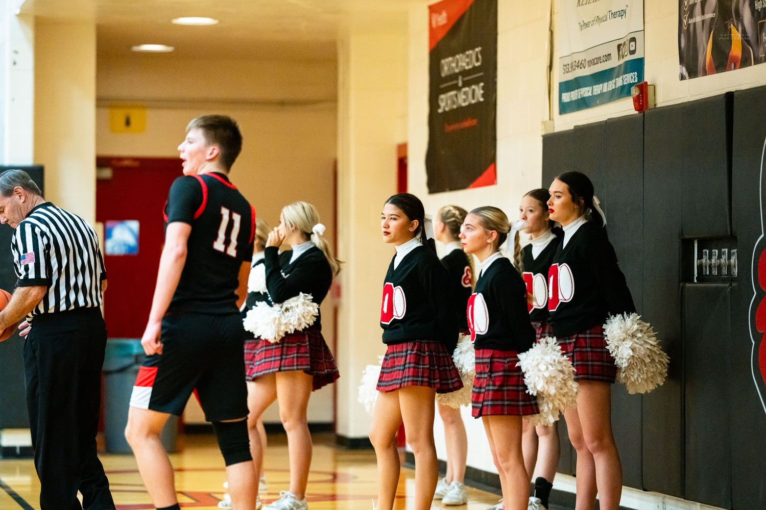 A referee and player in black number 11 jersey walk past cheerleaders with pom-poms along the gym wall.