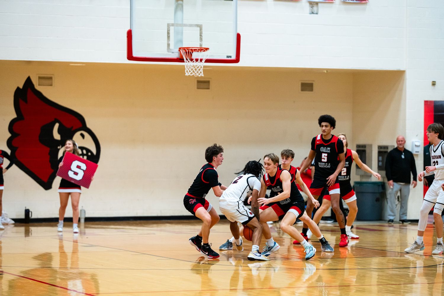 High school basketball players scramble for ball near Cardinals mascot logo during game.