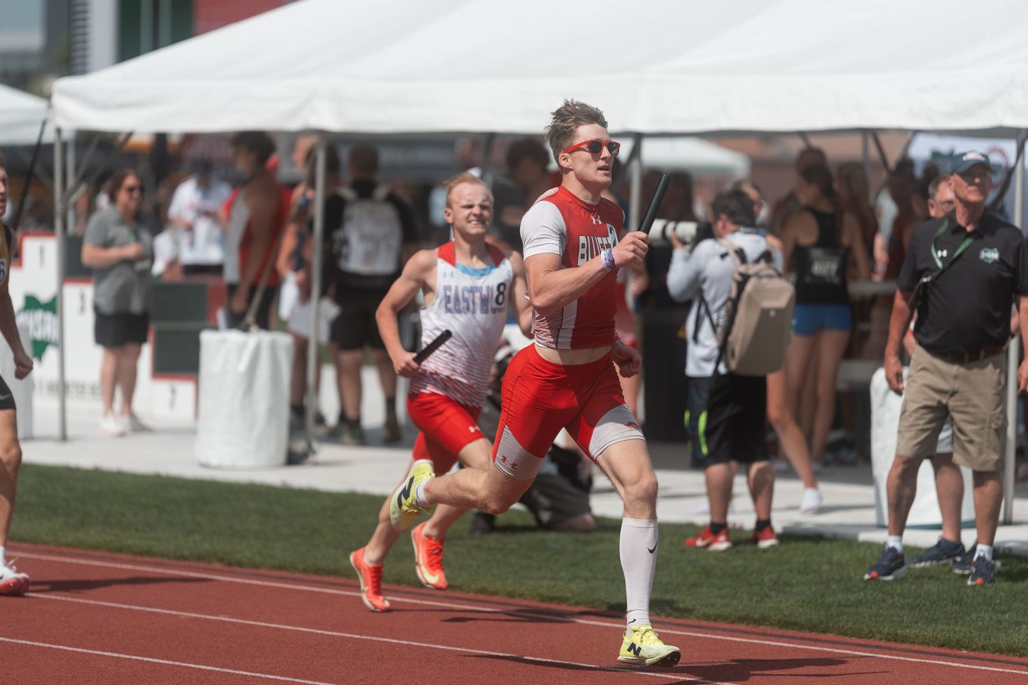 Two runners in red uniforms compete in a relay race on a red track at an outdoor championship meet.