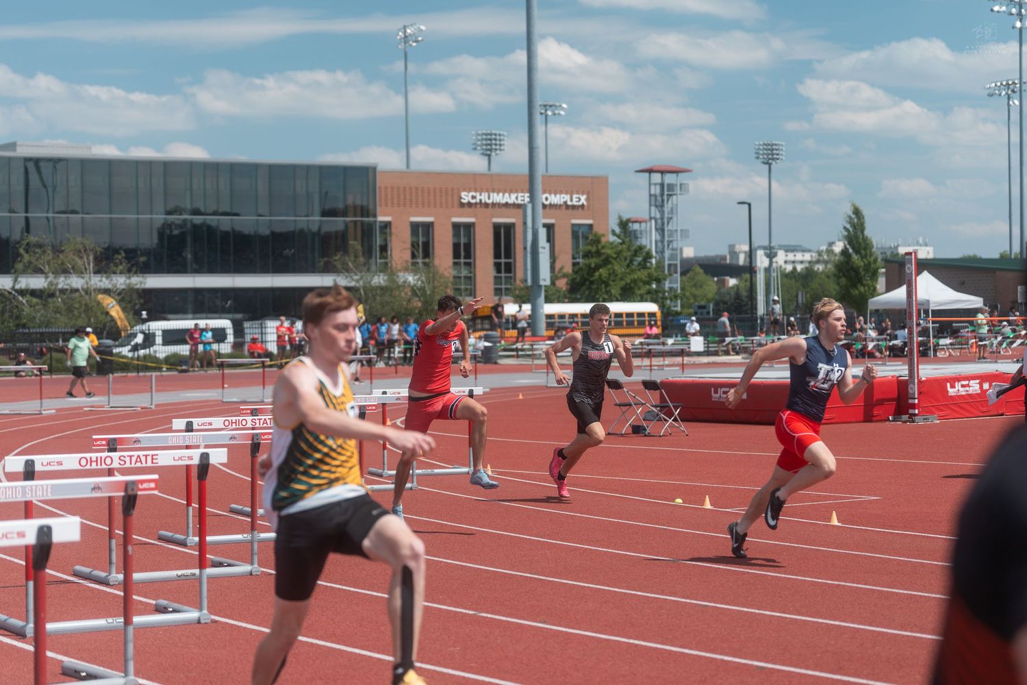 Young male athletes compete in a hurdles race at an outdoor track meet on a sunny day.