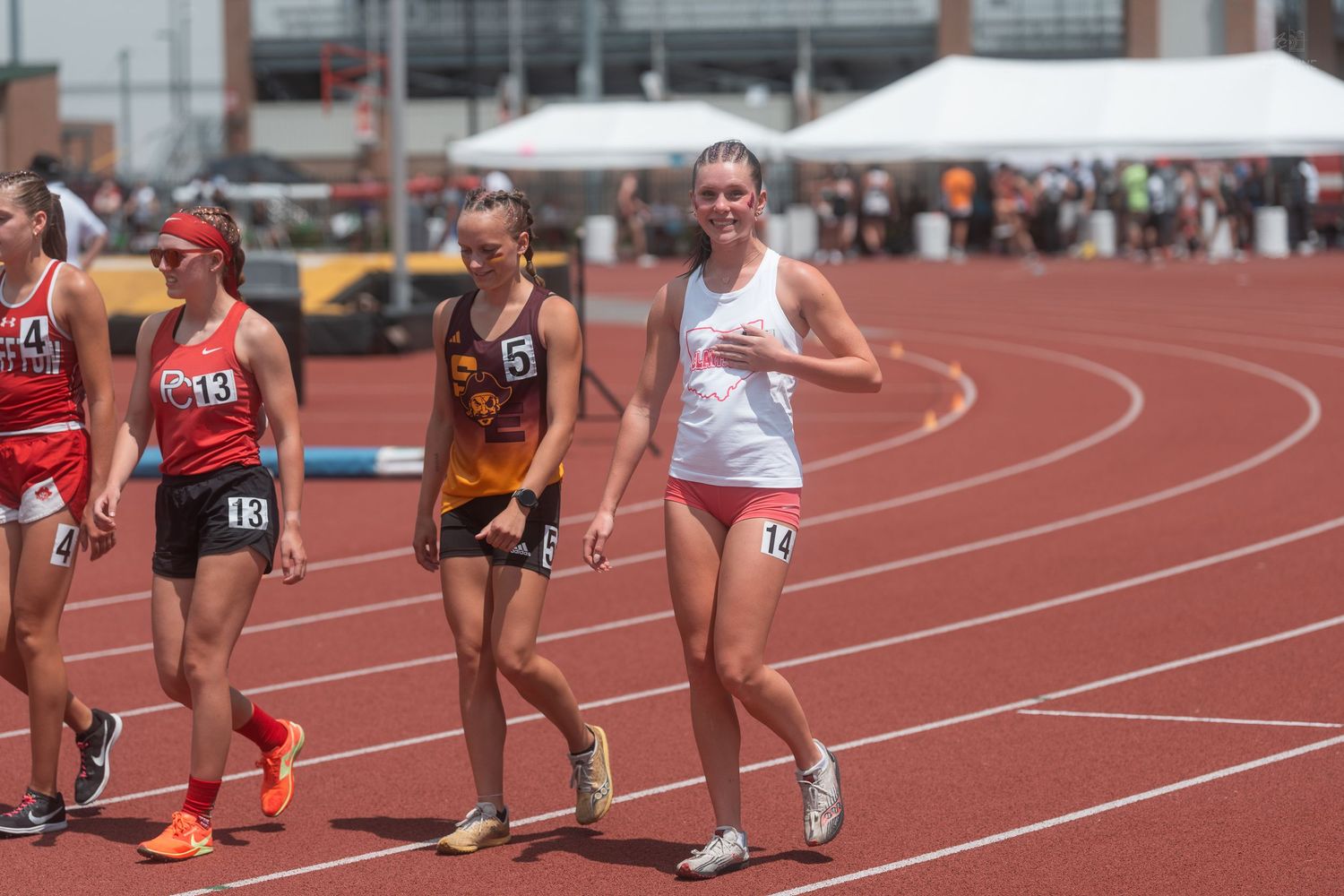 Female high school track runners in numbered uniforms competing on red outdoor track on a sunny day.