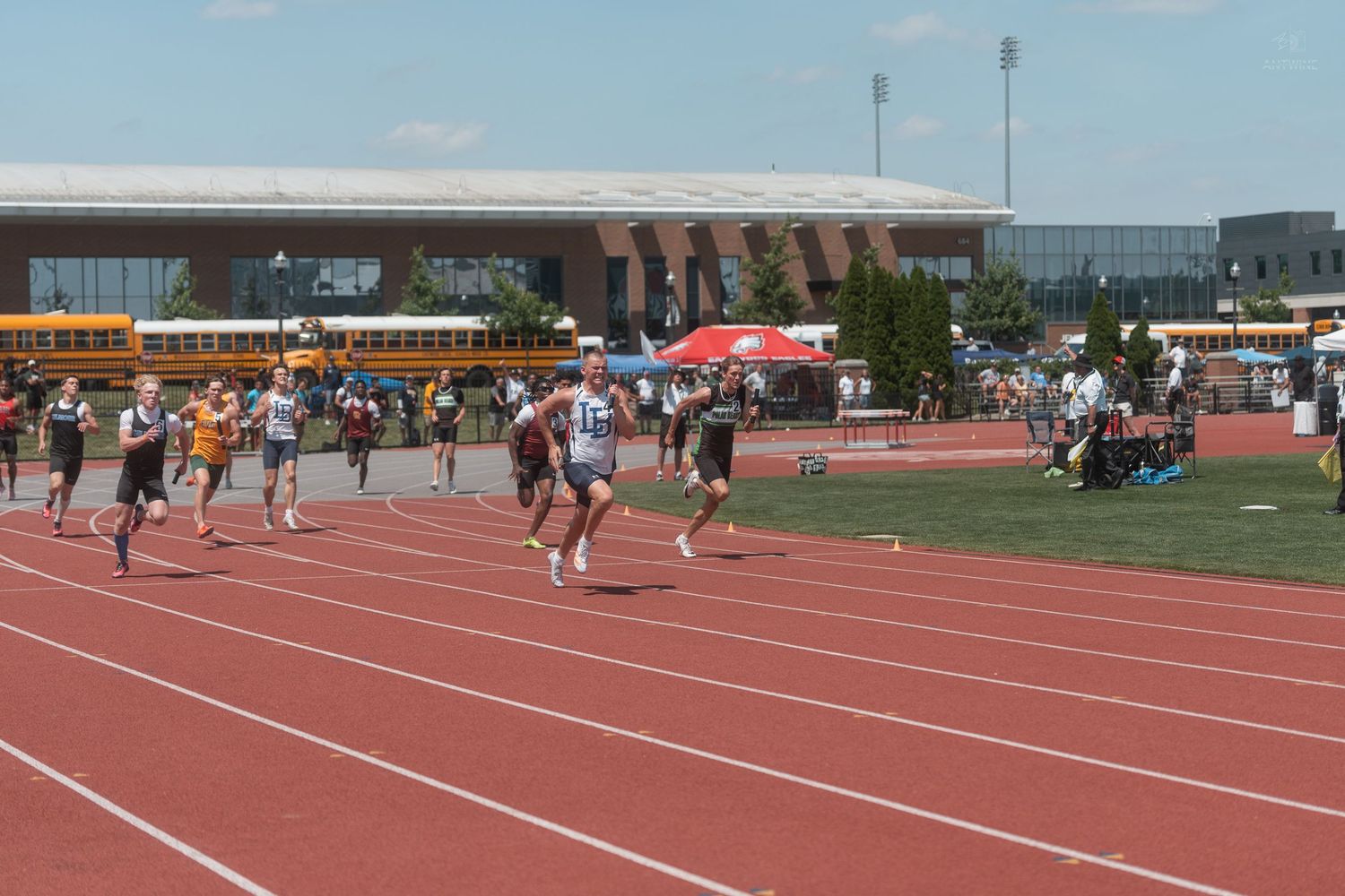 Athletes compete in a track race at an outdoor stadium with school buses and a large building visible in the background.