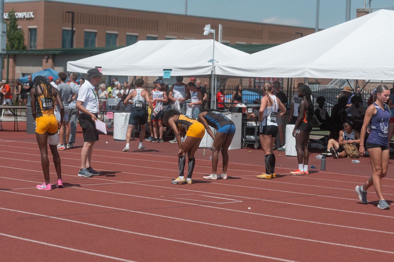 Female track athletes in gold uniforms prepare at the starting line during an outdoor track meet.
