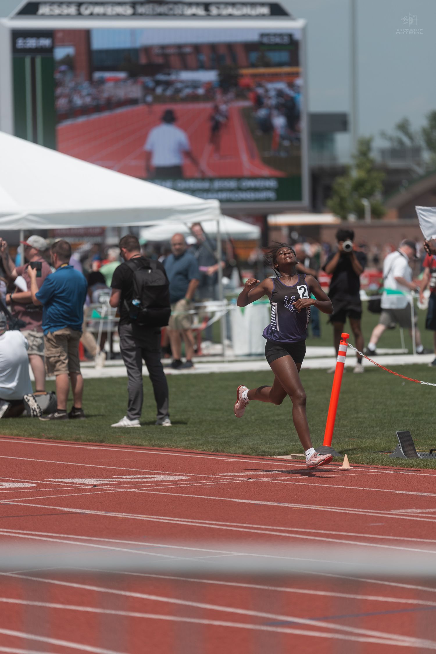 Female athlete in dark uniform crosses finish line at Jesse Owens Memorial Stadium during a championship track meet.