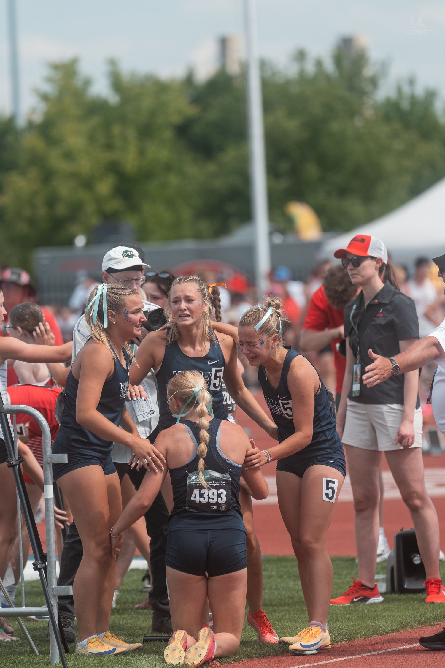 Female track athletes in dark uniforms huddle together celebrating at an outdoor track and field competition.