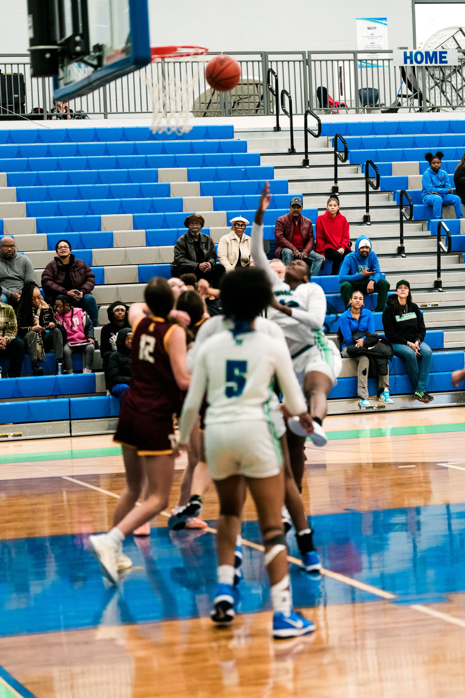Female basketball players compete near the basket during a high school girls basketball game in a blue gymnasium.