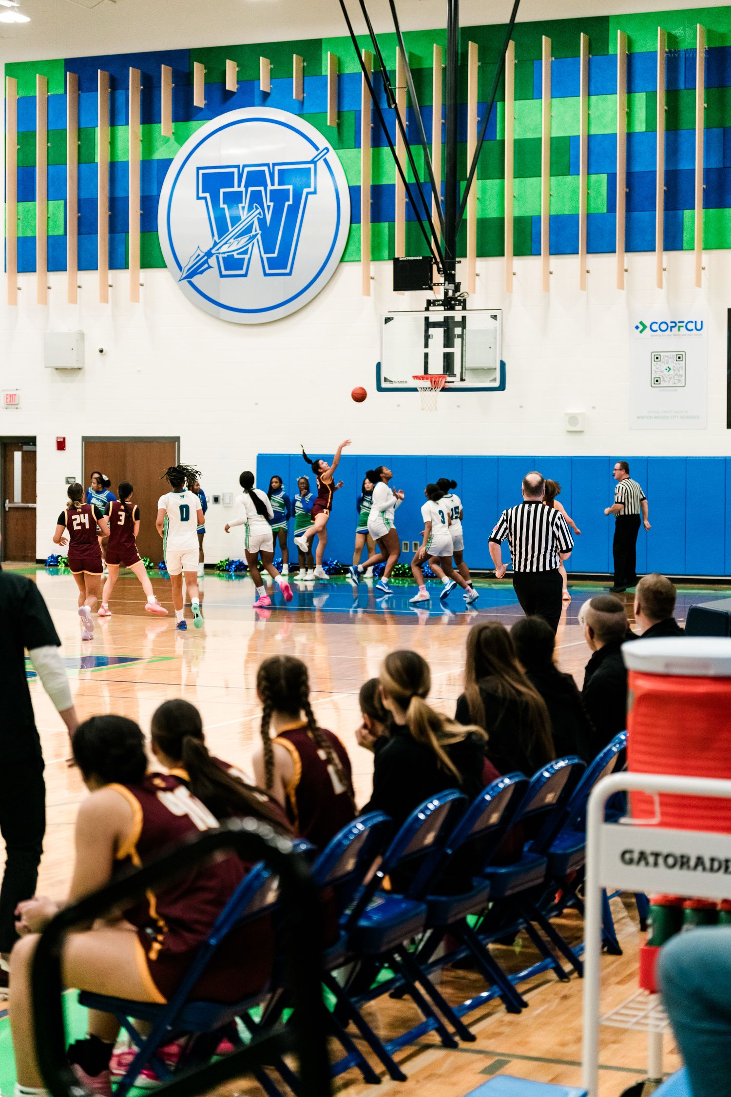 Wide-angle view of a girls high school basketball game inside Winton Woods gymnasium with spectators watching.