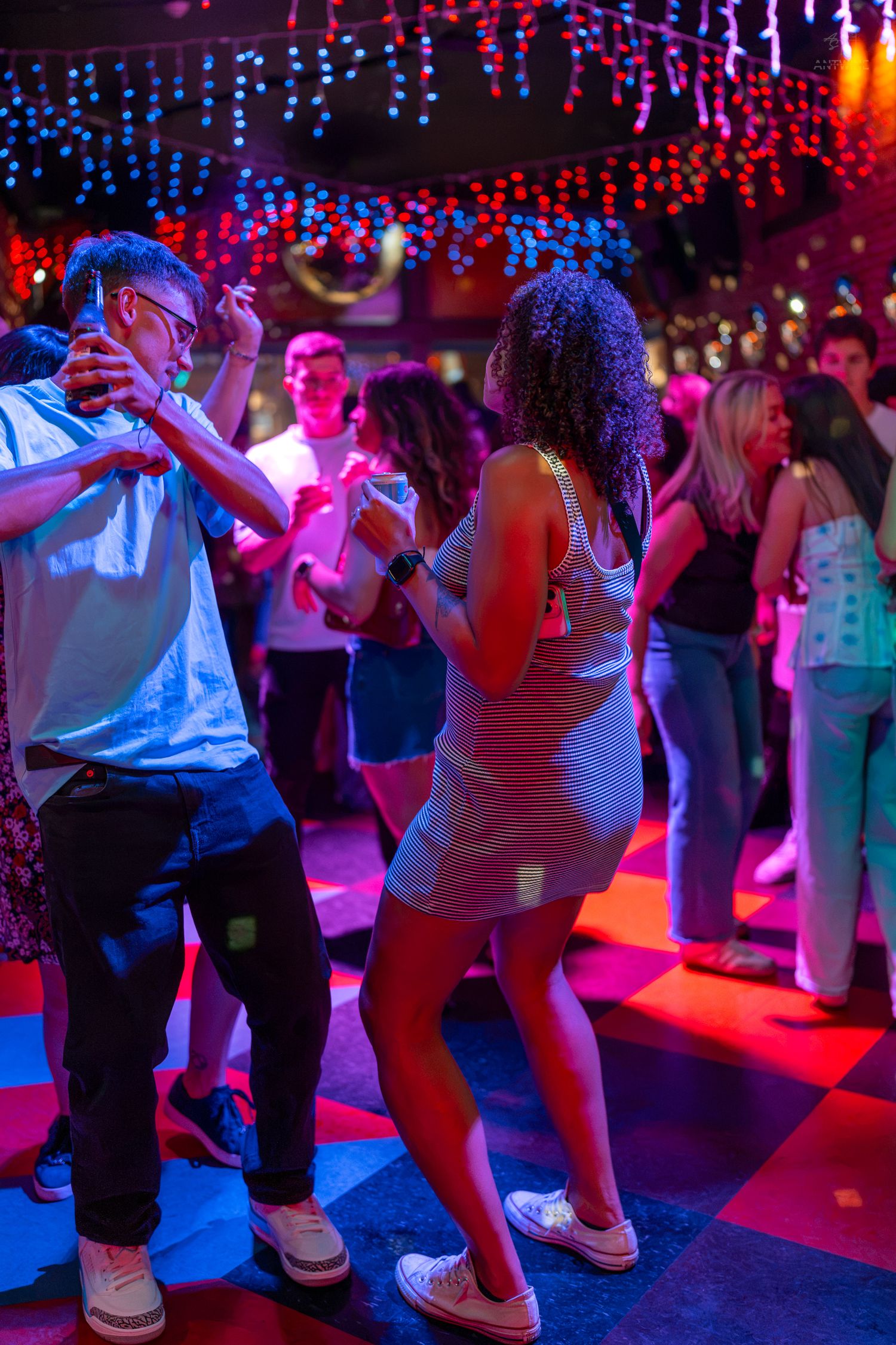 Couples dancing on a colorful checkered floor in a vibrant nightclub with red and blue lights.