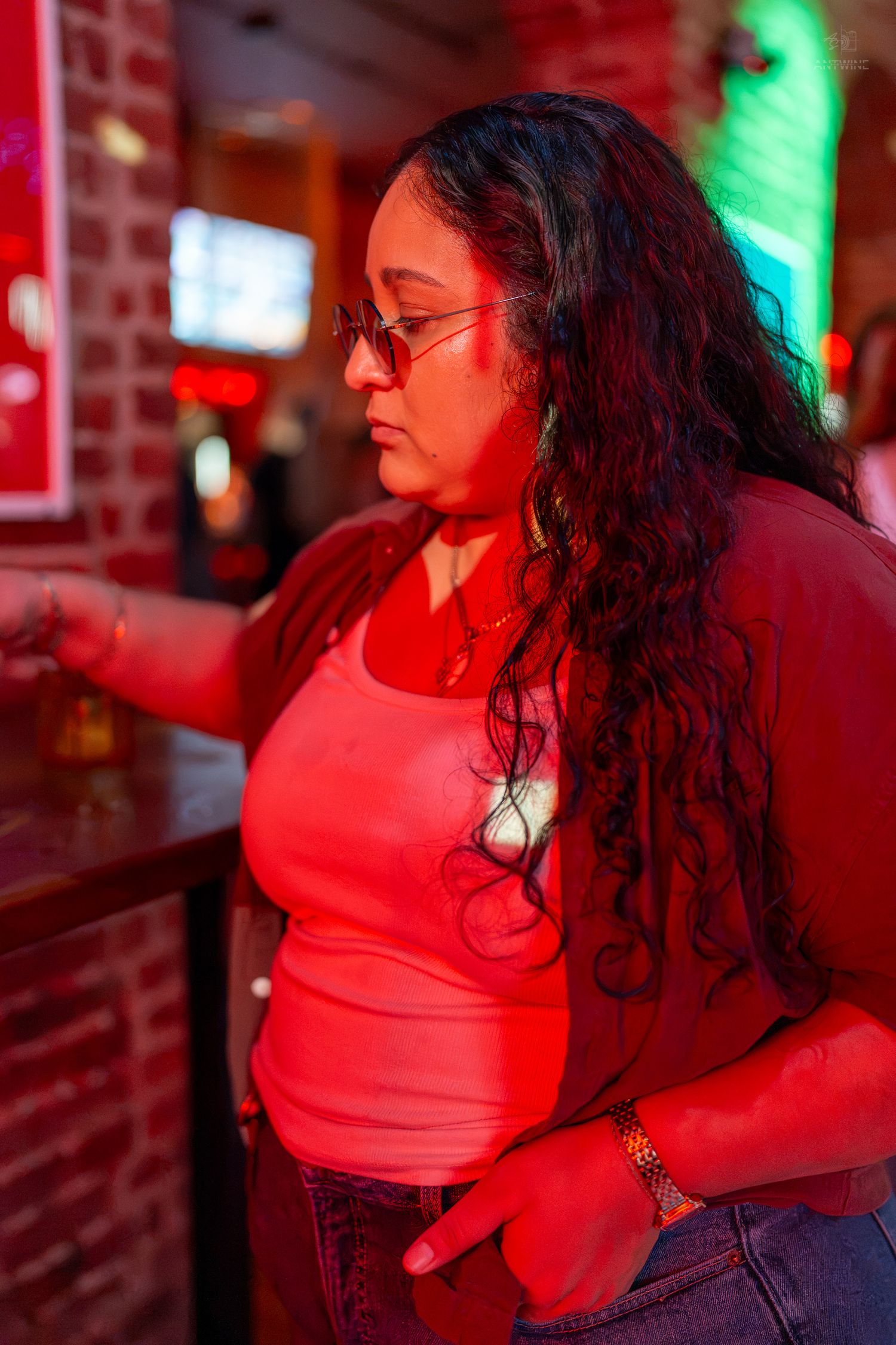 Woman with long curly hair and glasses standing at a bar under red neon lights.