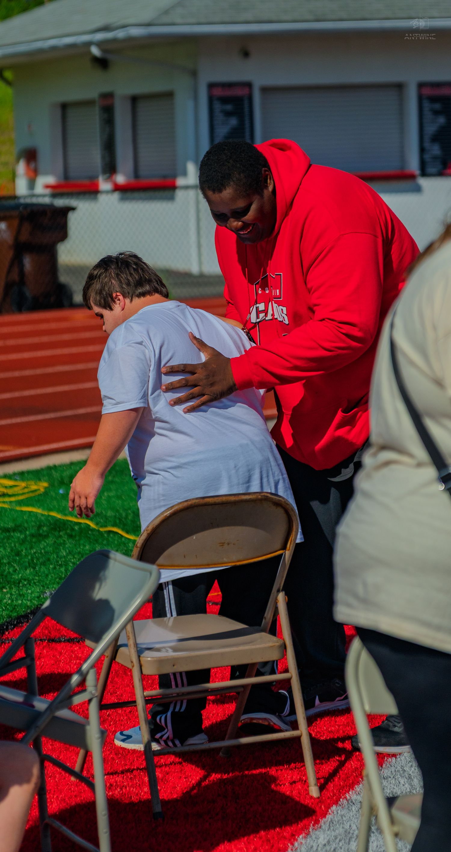 A person in a red hoodie assists another seated on a folding chair on a red track field outdoors.