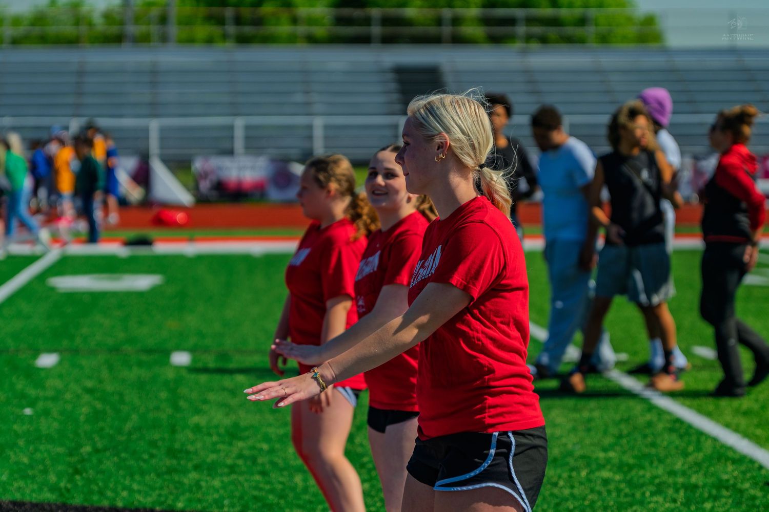 Young women in red shirts compete in a track and field event on a green turf stadium.