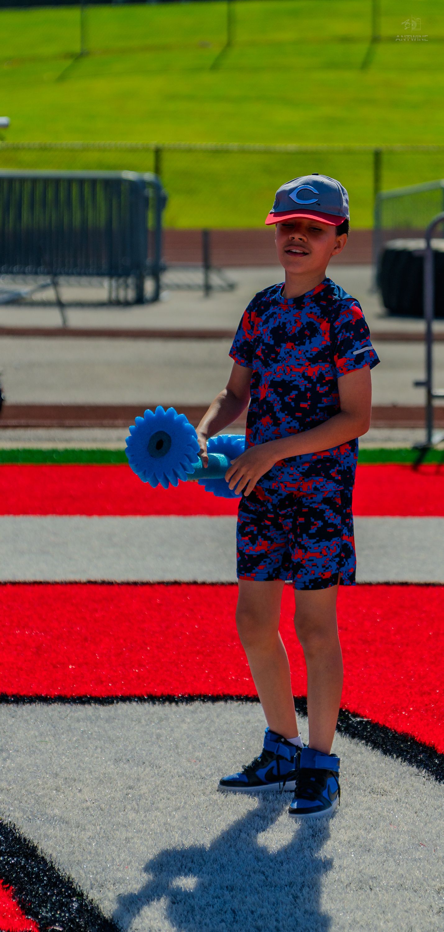 Boy in Cincinnati Reds cap holding blue flower toy on red turf field.