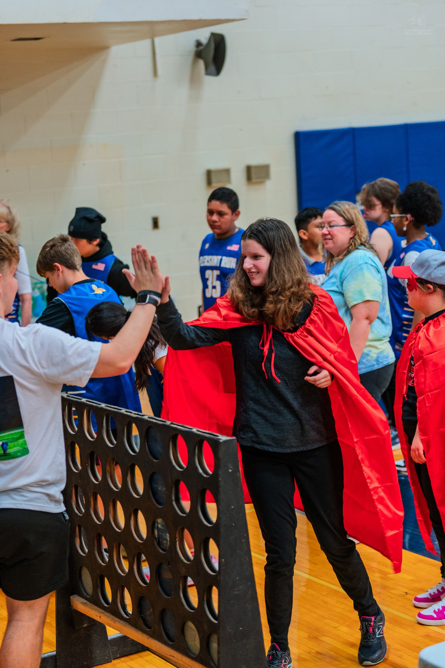 Students high-five during a gym activity featuring a giant Connect Four game, with participants wearing superhero capes.