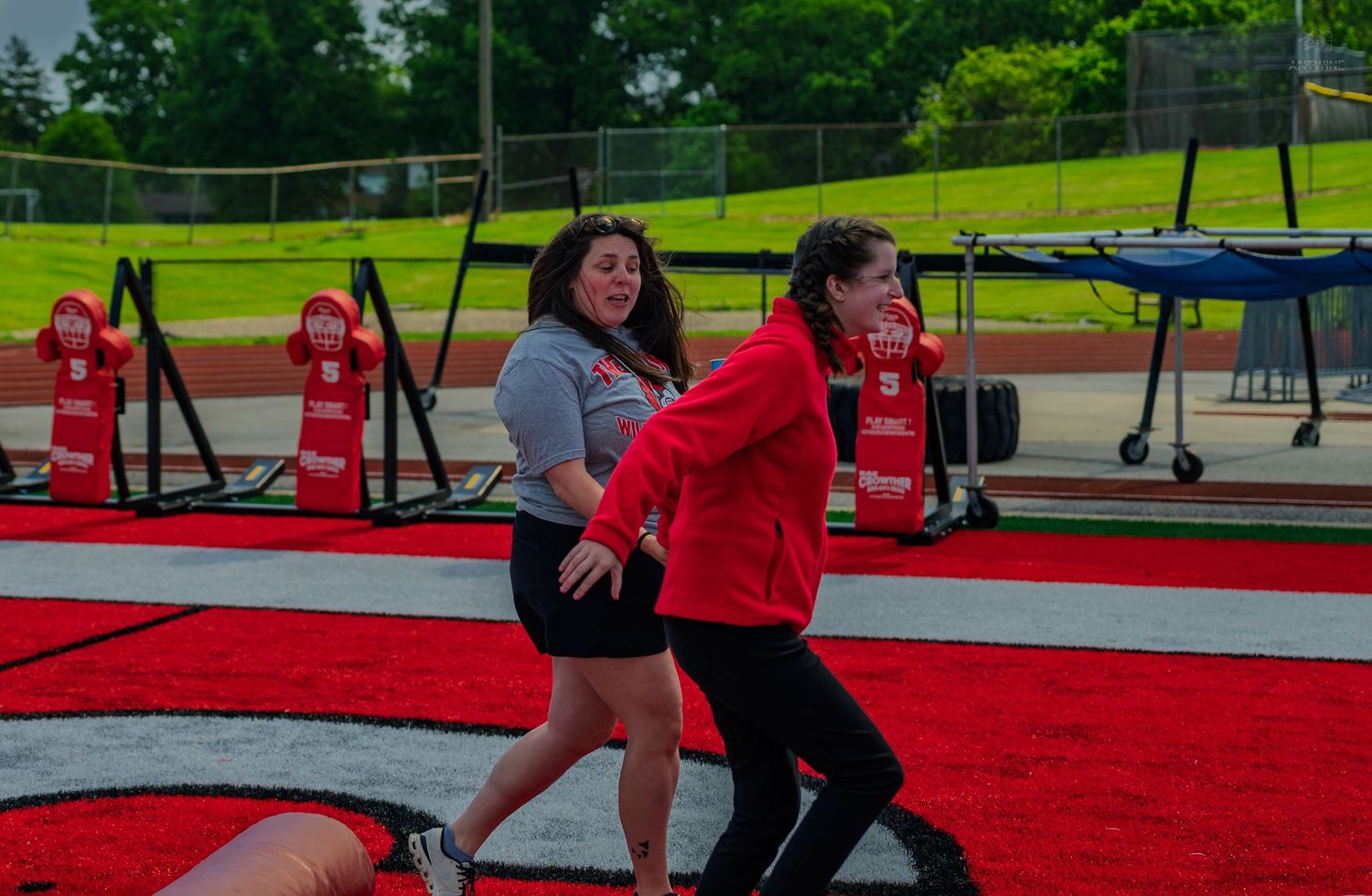 Two women walking together on a red track at an outdoor athletic field during a cloudy day.