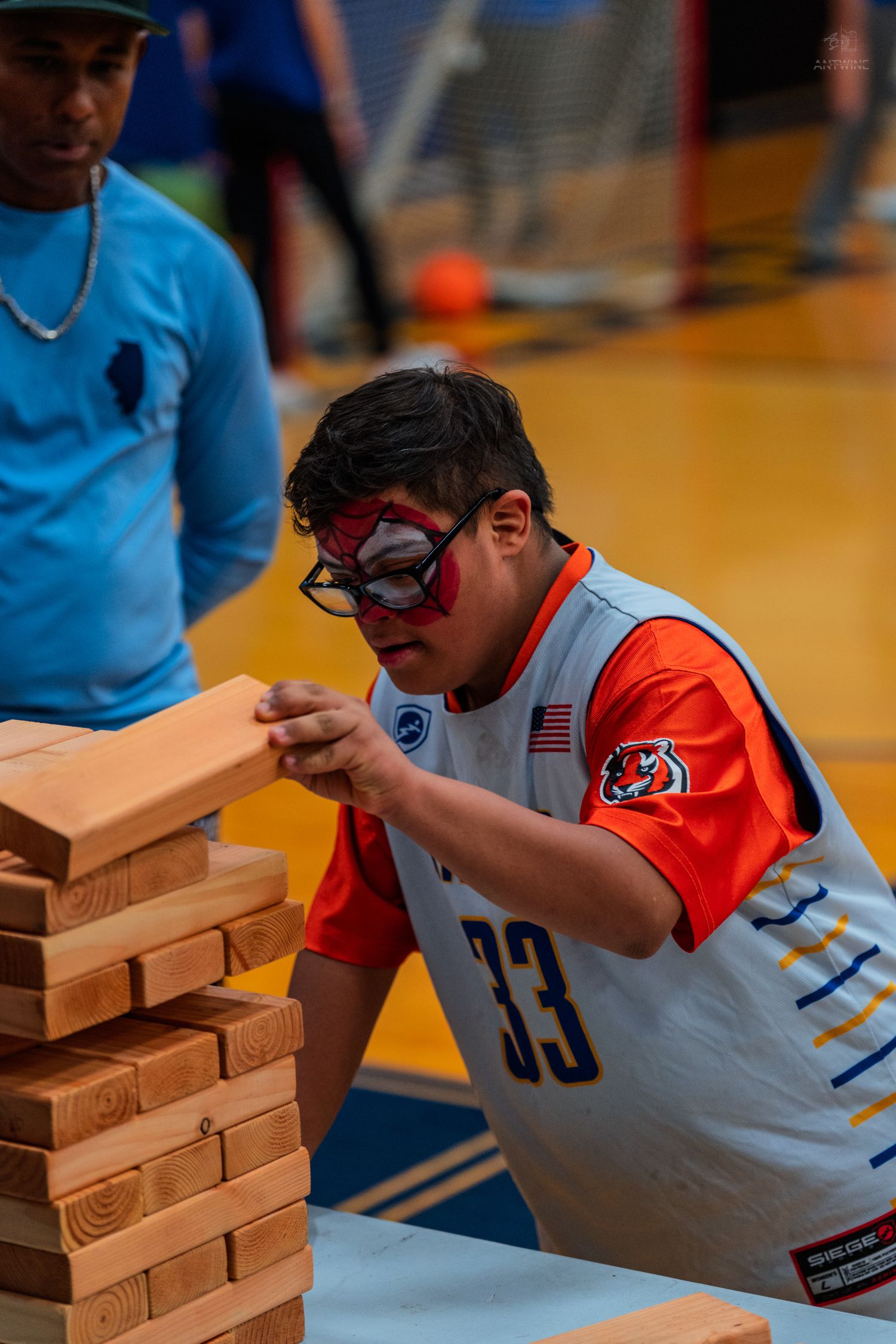 Young boy wearing Spider-Man mask and jersey #33 plays giant Jenga at an indoor recreational event.