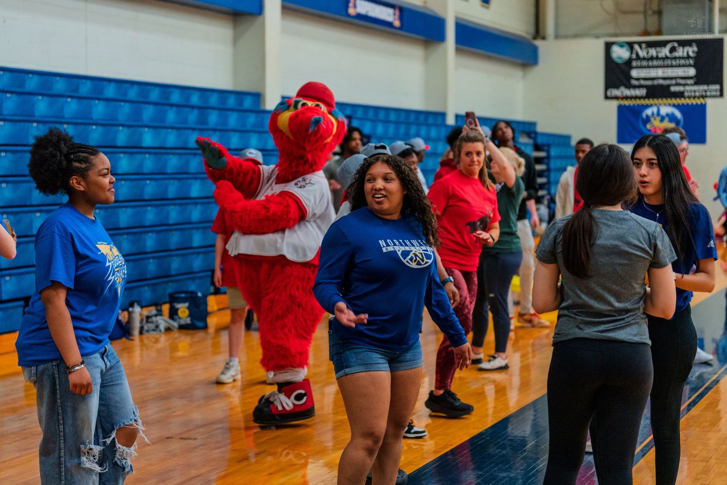 Students dance with a red cardinal mascot in a school gymnasium during a lively campus event.