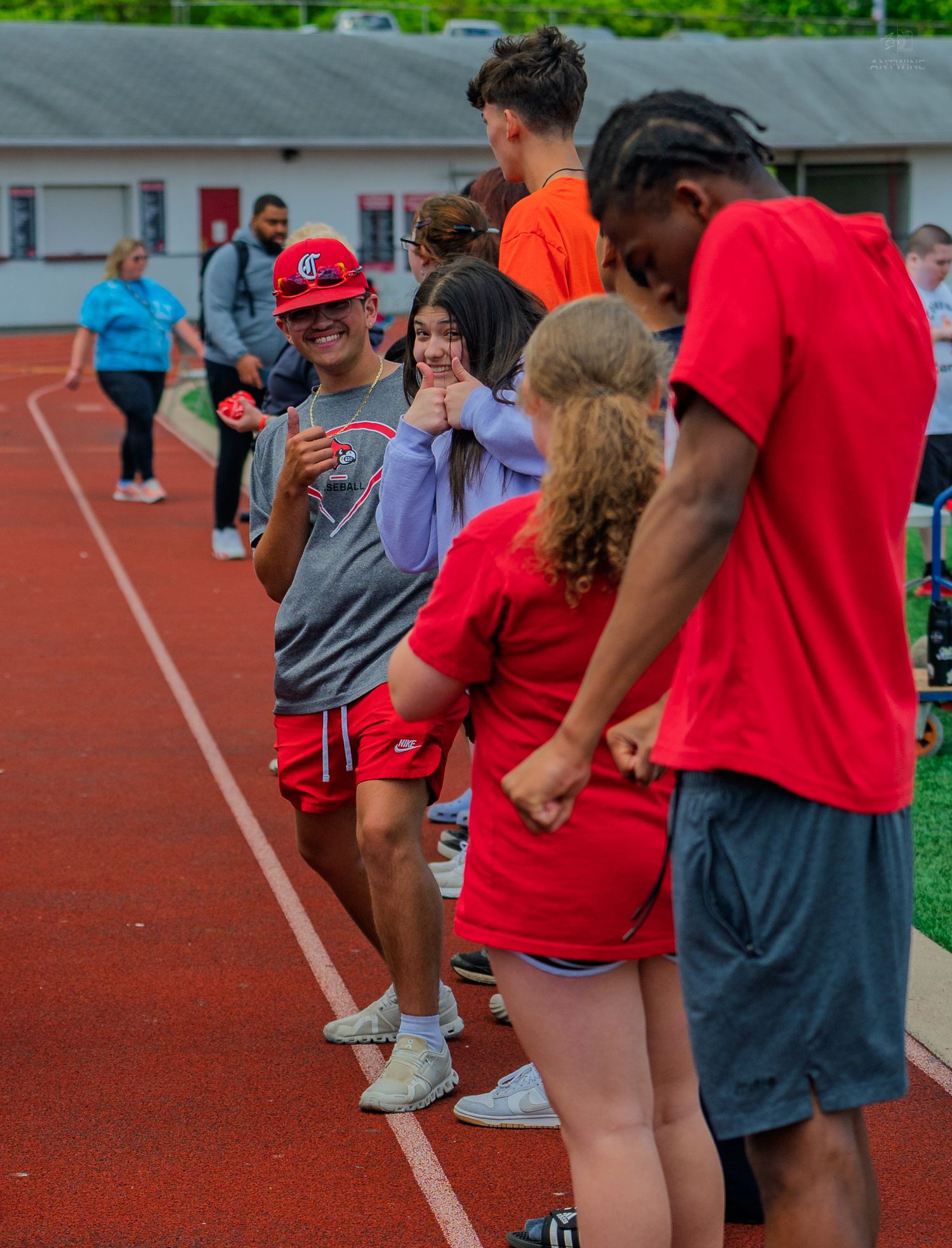 Students in red uniforms laugh and interact on a running track during an outdoor school sports event on a sunny day.