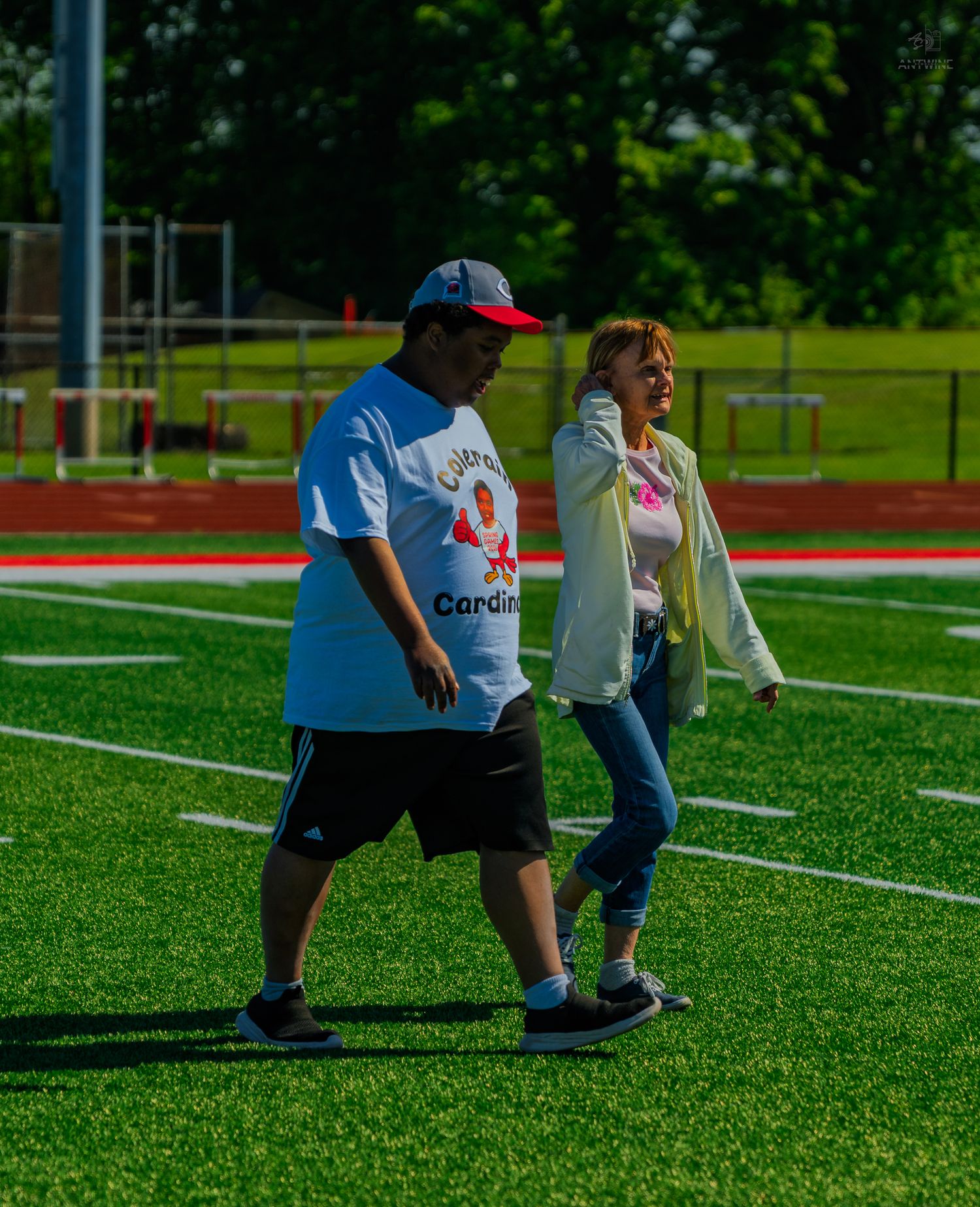 Two people walking on a green turf football field, man wearing Cardinals jersey and red cap.