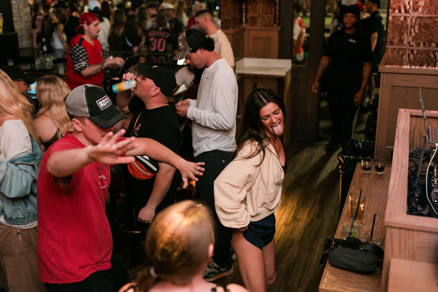 A cheerful woman in a cream hoodie dances energetically among a lively crowd inside a busy bar.