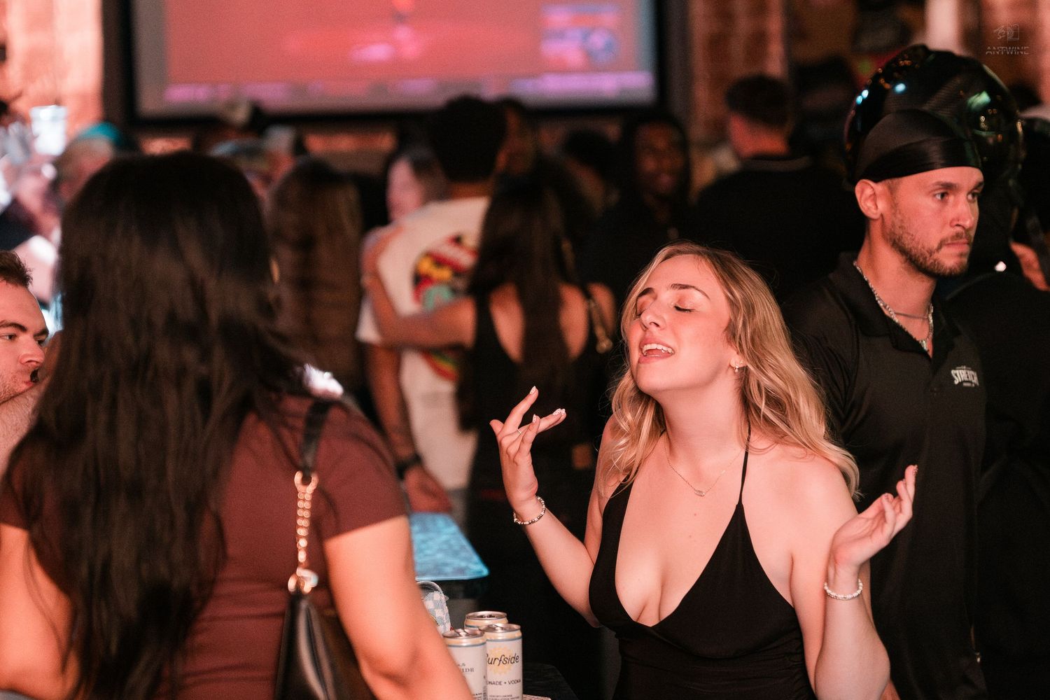Smiling blonde woman gestures expressively while chatting with friends at a lively bar venue.