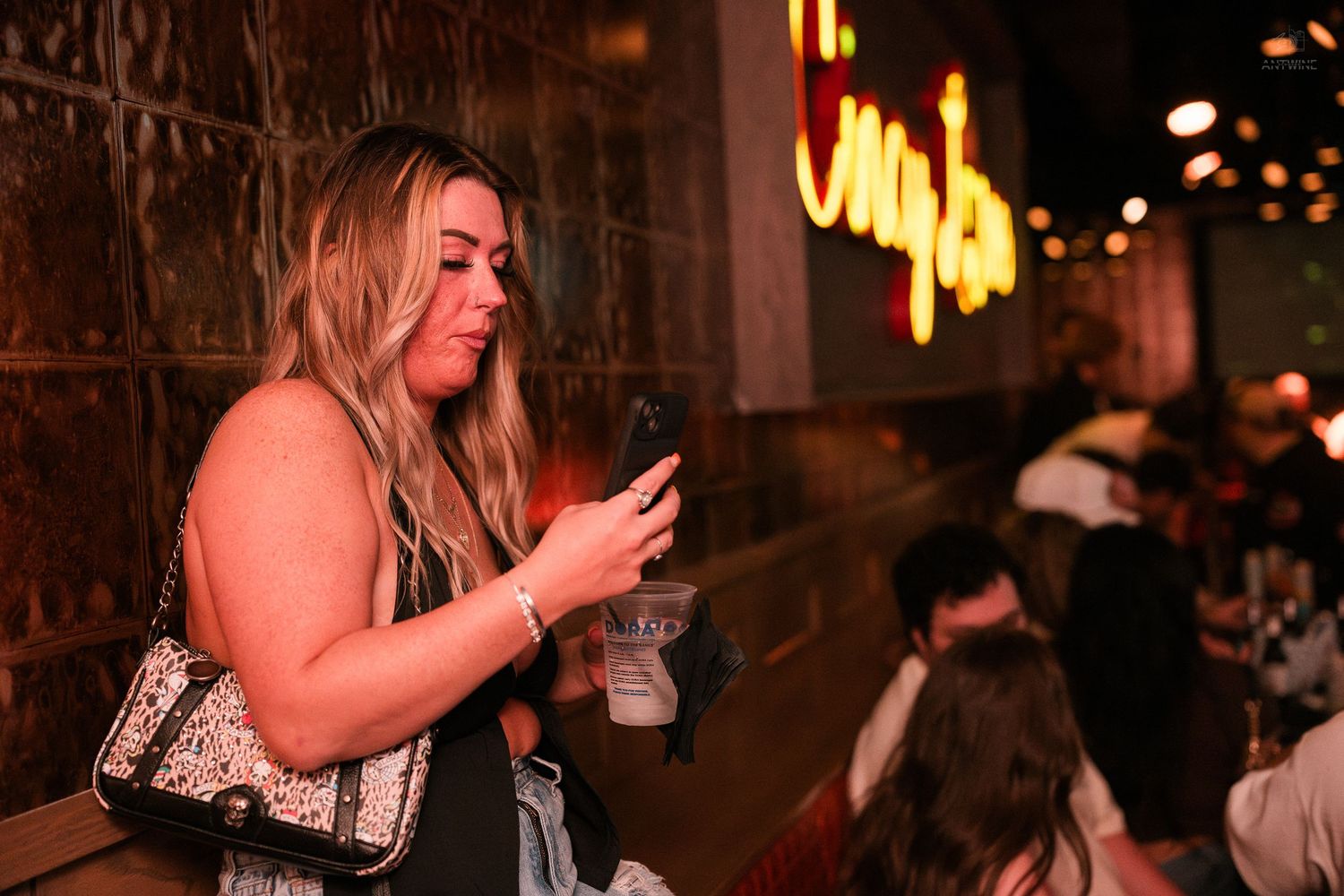 Blonde woman checks her phone while holding a drink at a neon-lit bar or nightclub venue.