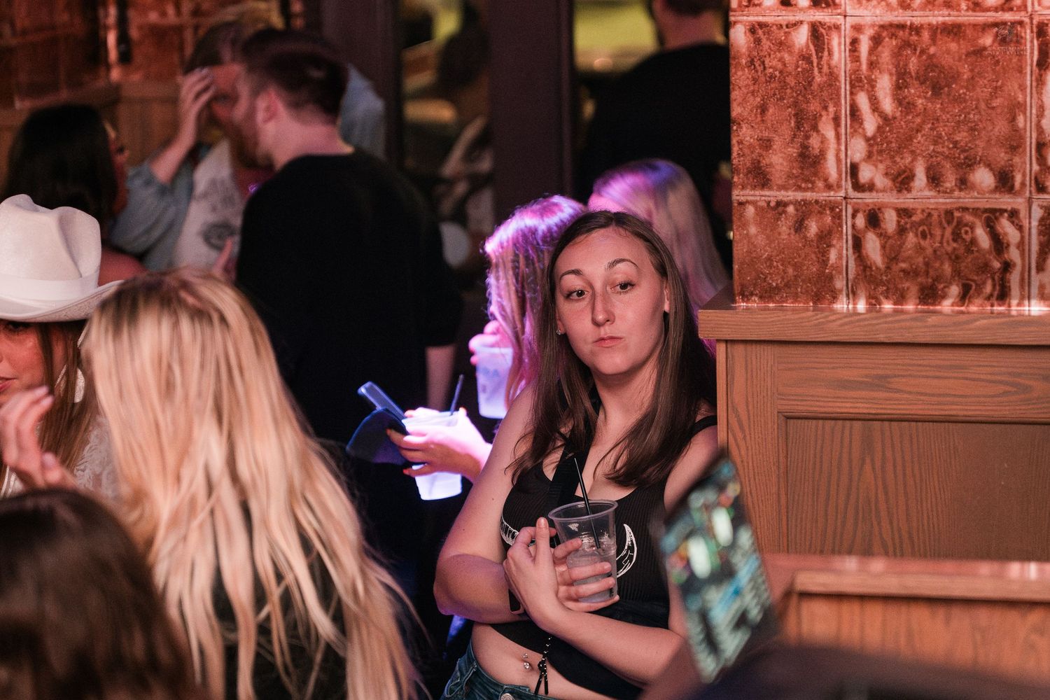 Young woman holding a drink looks back at the camera in a dimly lit bar setting.