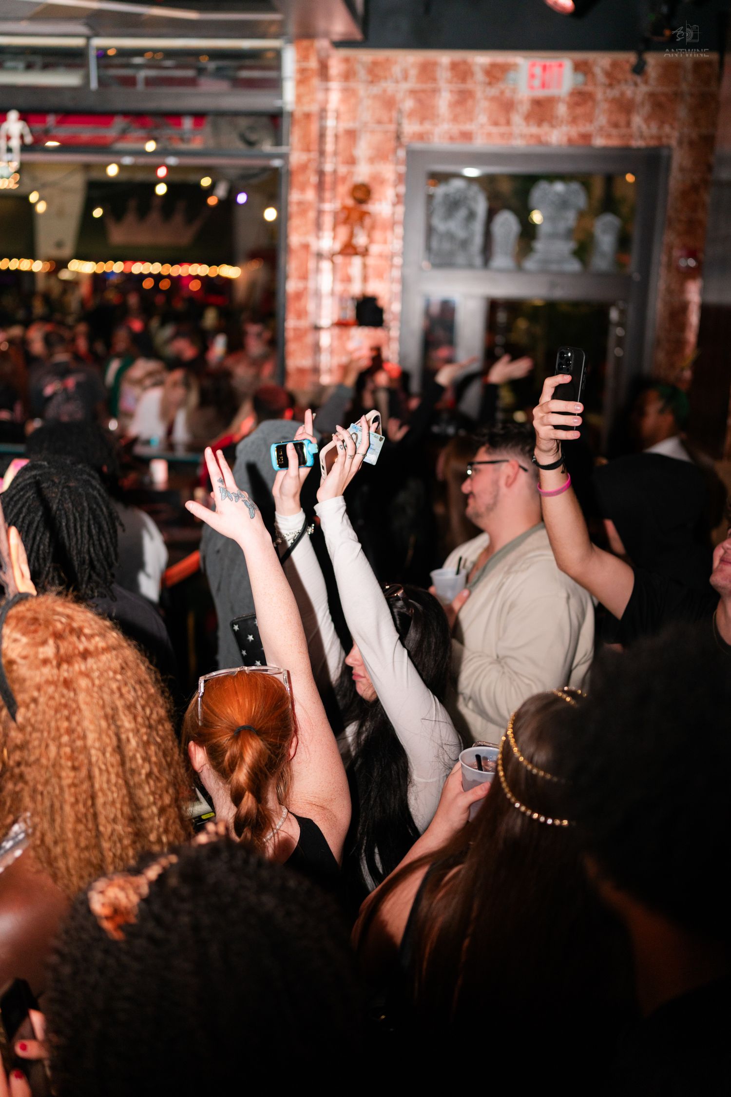 Energetic Halloween party crowd with raised hands and phones in warmly lit brick venue.