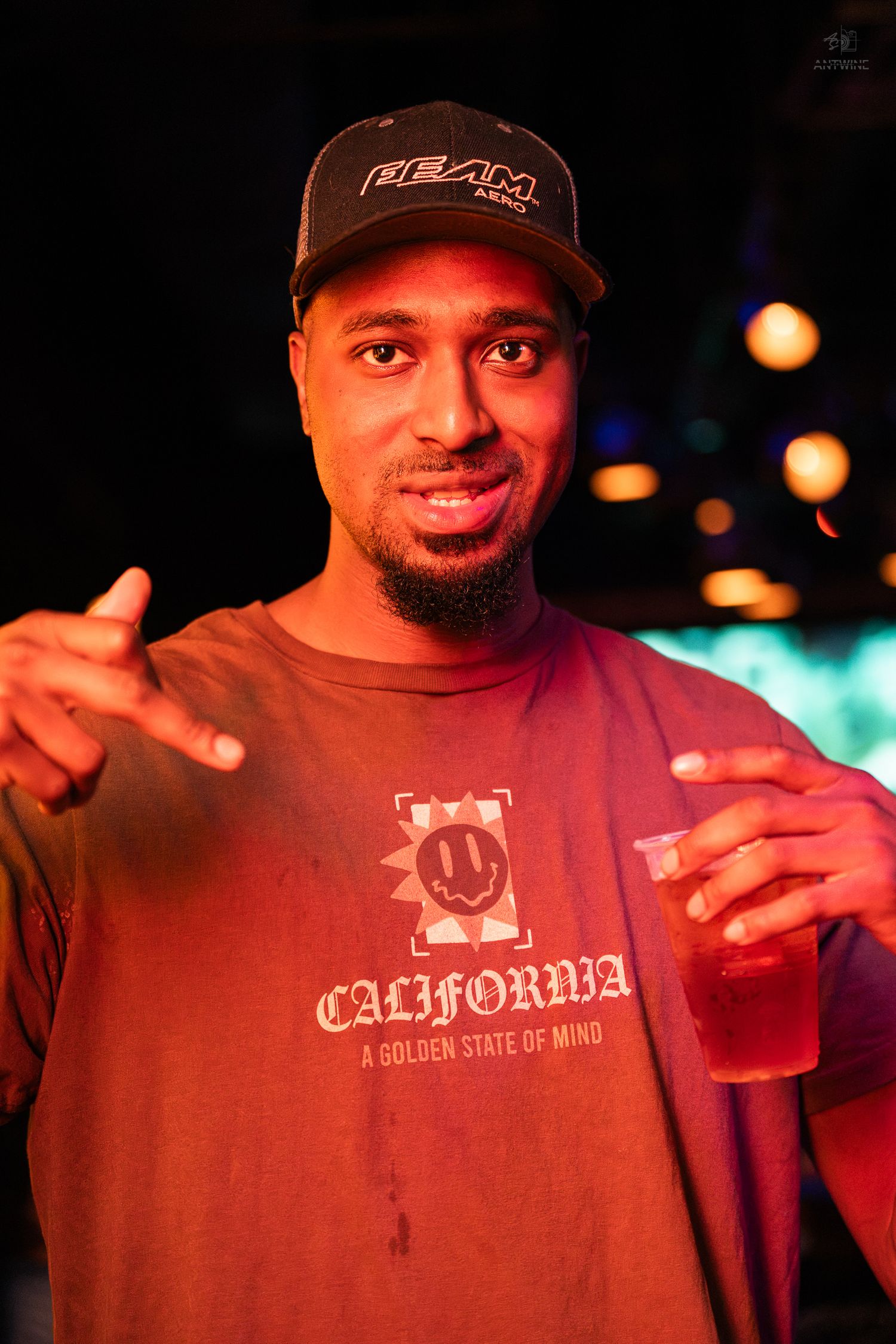 A man in a California t-shirt and cap holds a red drink at a nightclub event.