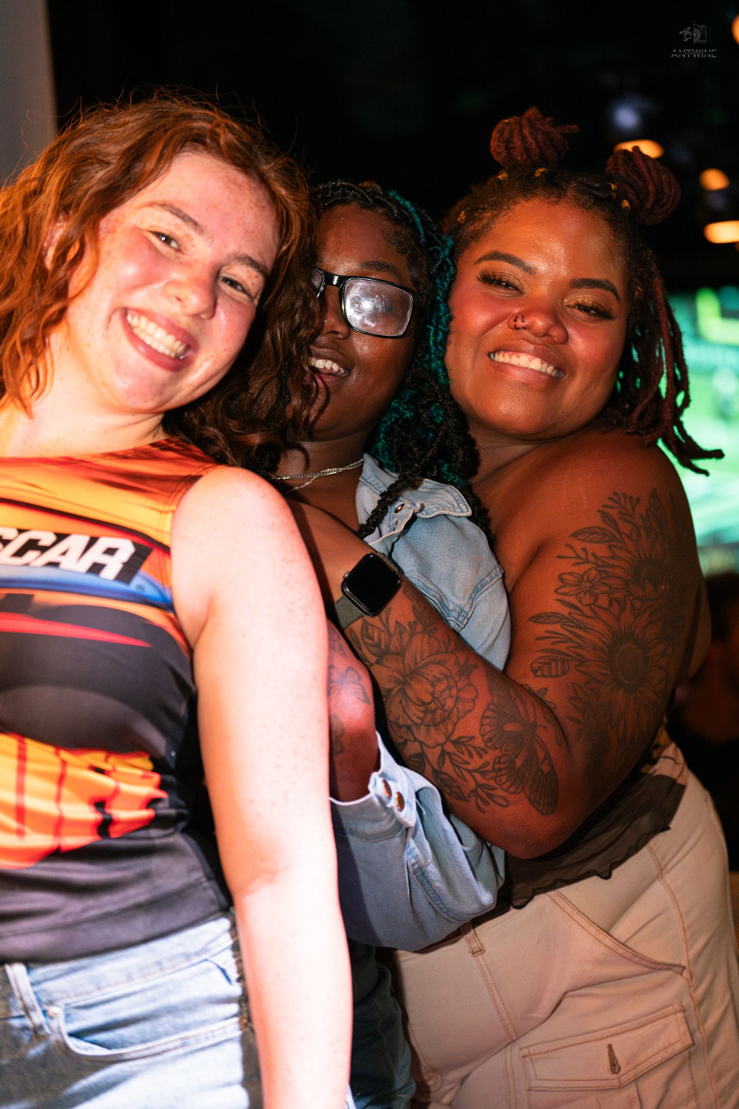 Three smiling women posing together at a nightclub, one wearing a NASCAR top and another with colorful hair and tattoos.