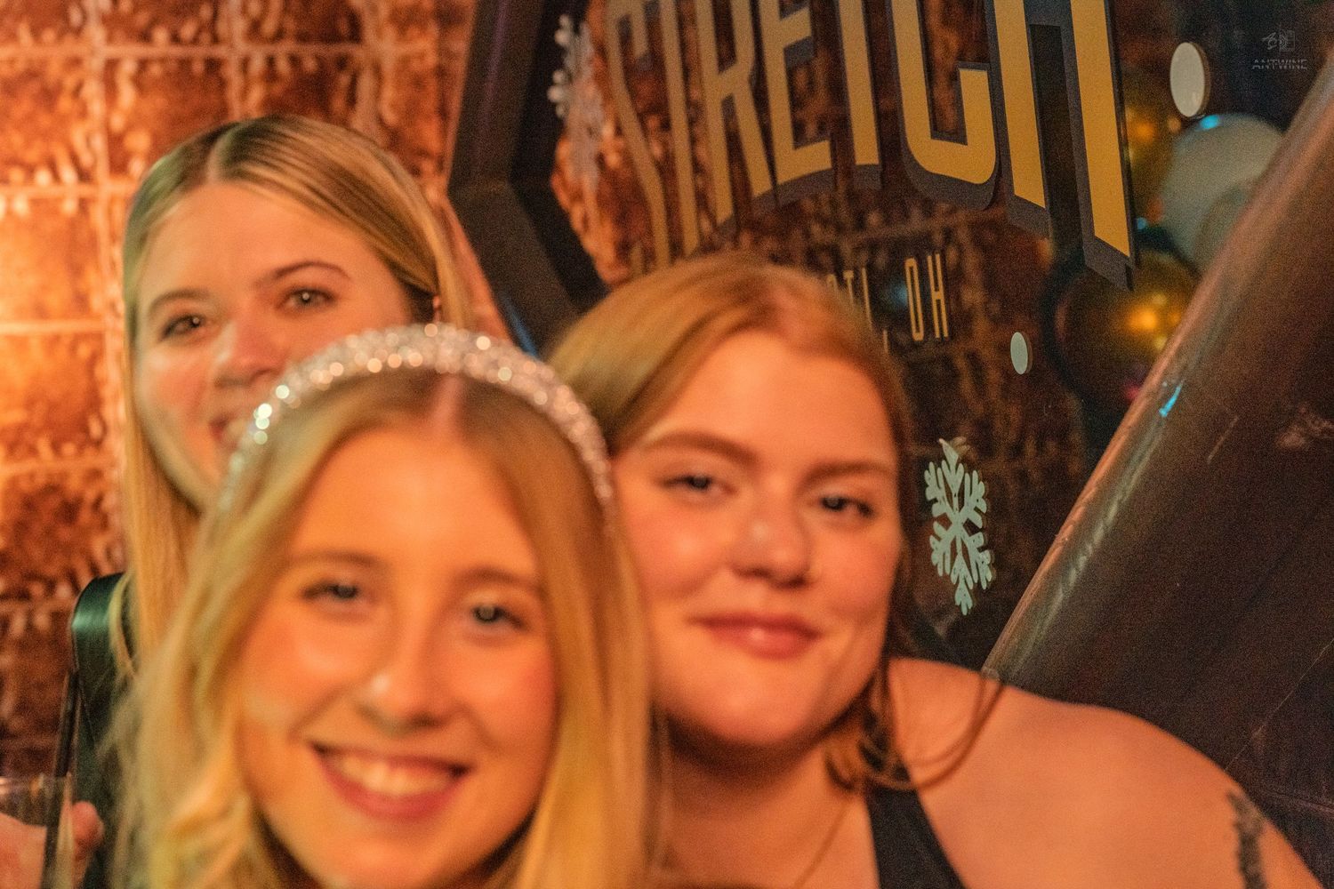 Three women smiling together near a Stretch bar sign at a party.