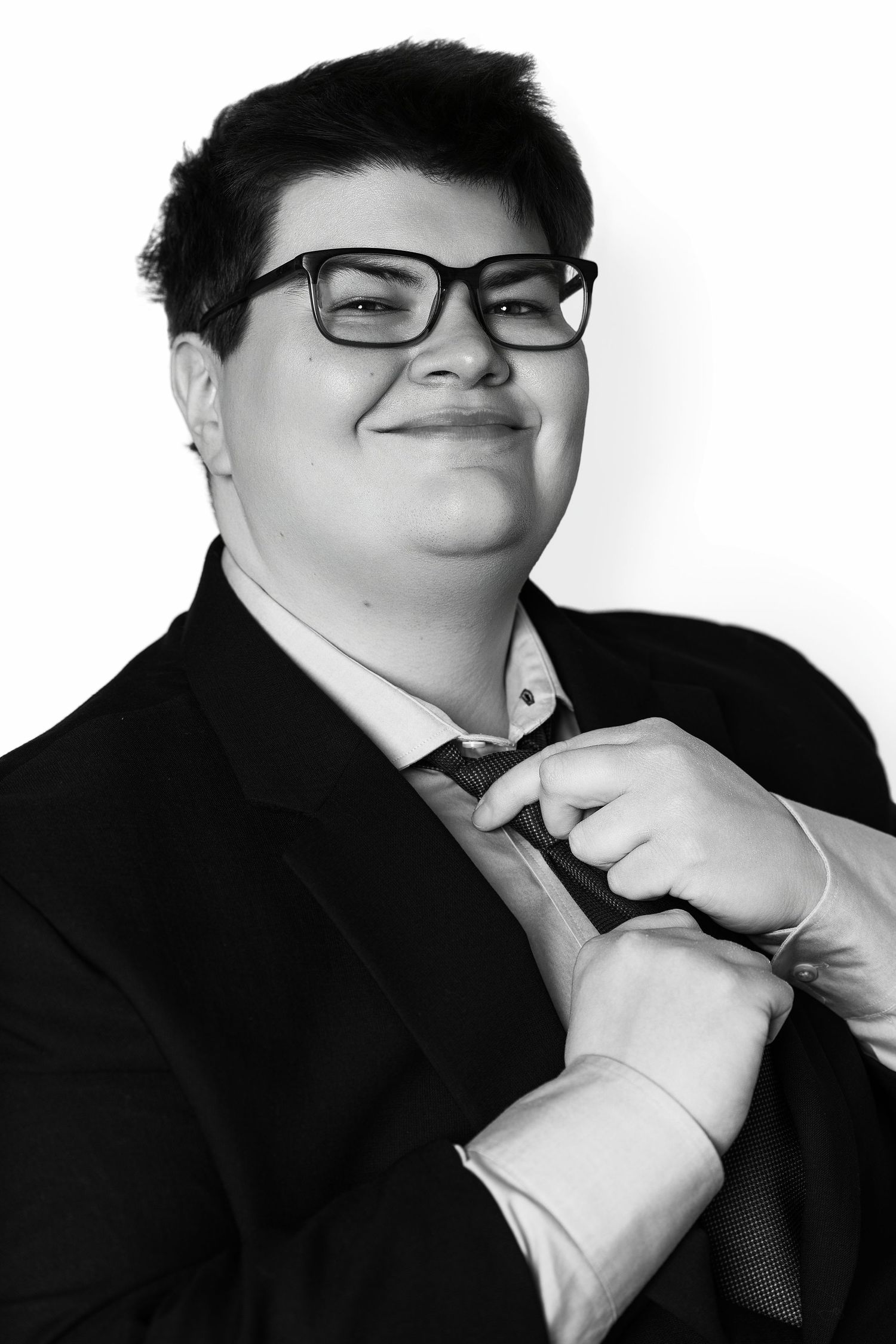 Black and white portrait of smiling young person in suit and glasses adjusting tie.