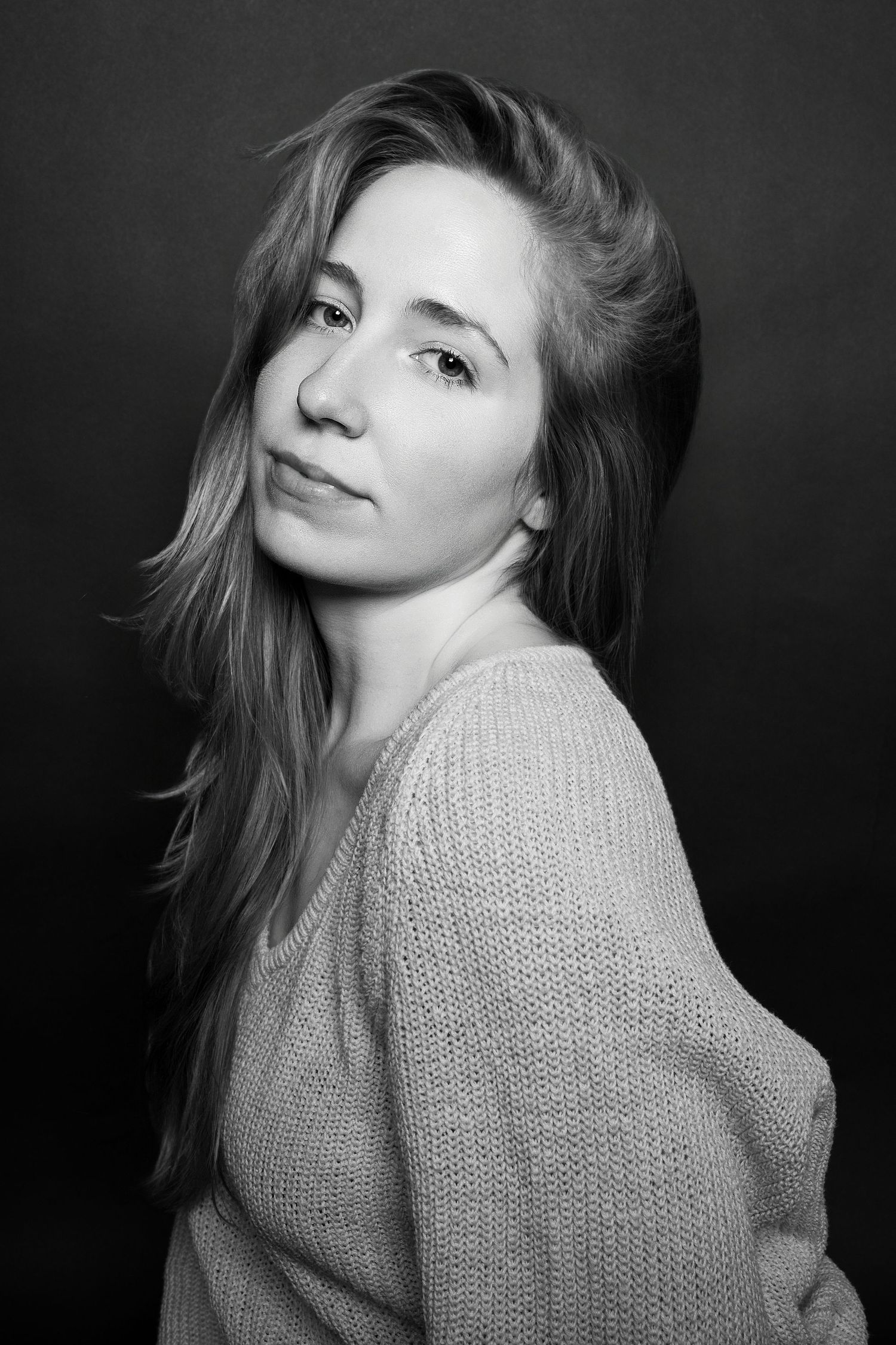 Black and white portrait of a woman with flowing hair in a knit sweater, looking over her shoulder.