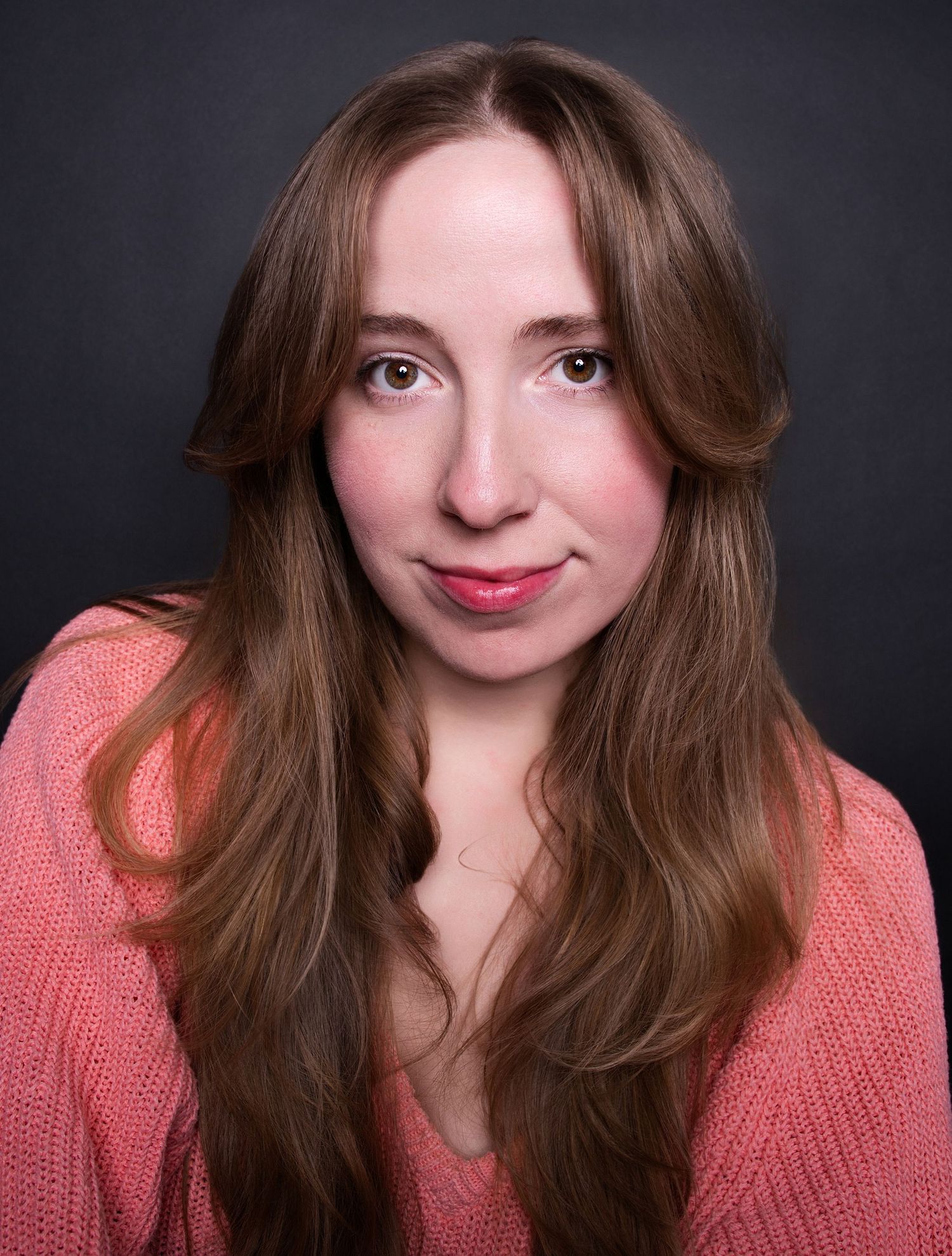 Young woman with long auburn hair smiling softly, wearing a pink knit sweater against a dark gray background.