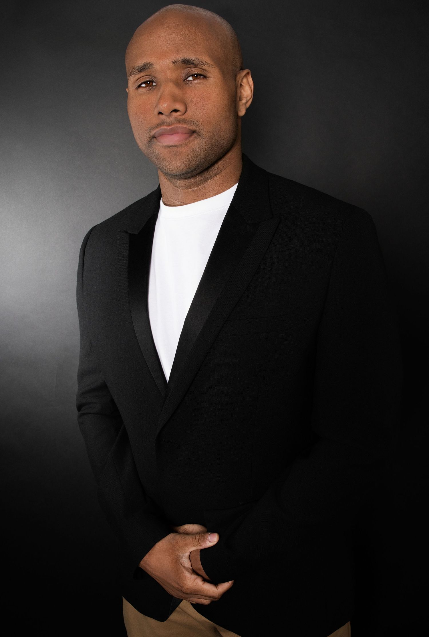 Bald Black man in black blazer and white shirt poses confidently against dark background.