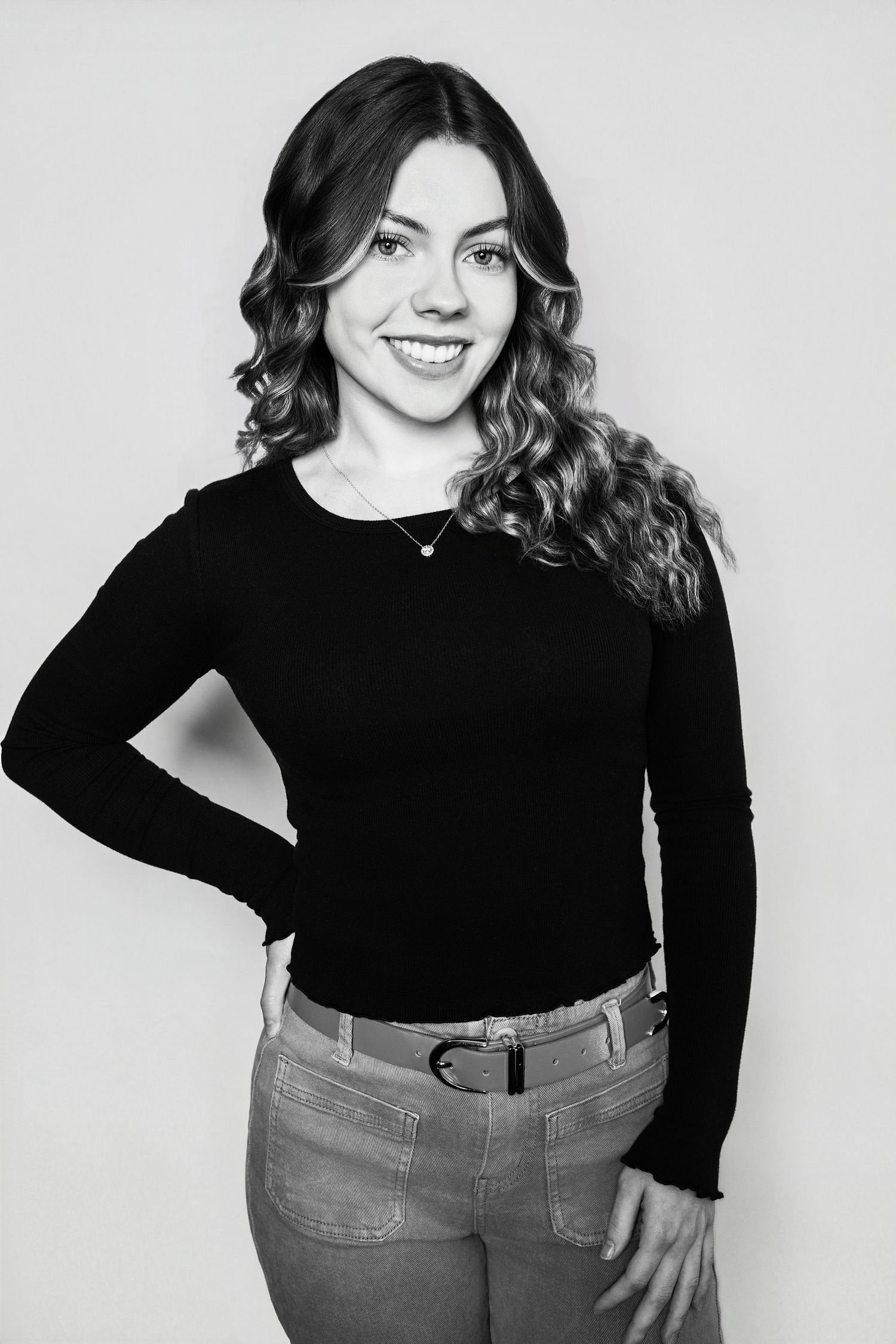 Smiling young woman in a black long-sleeve top and jeans poses confidently in a black and white photo.