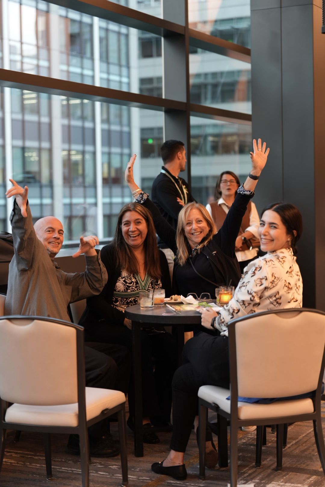 A group of smiling people raise their hands at a round table during a corporate event.