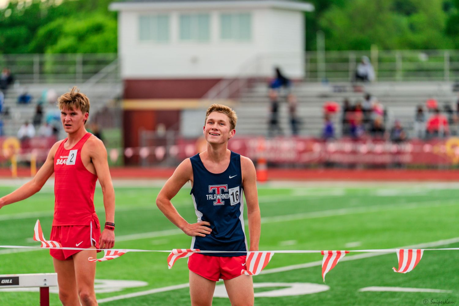 Two male track athletes in red uniforms stand near hurdles on a red track after a race.