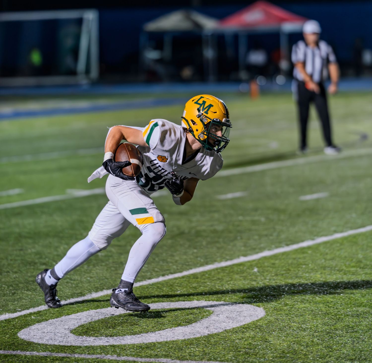 High school football player in white and gold uniform carries the ball along the sideline at night.