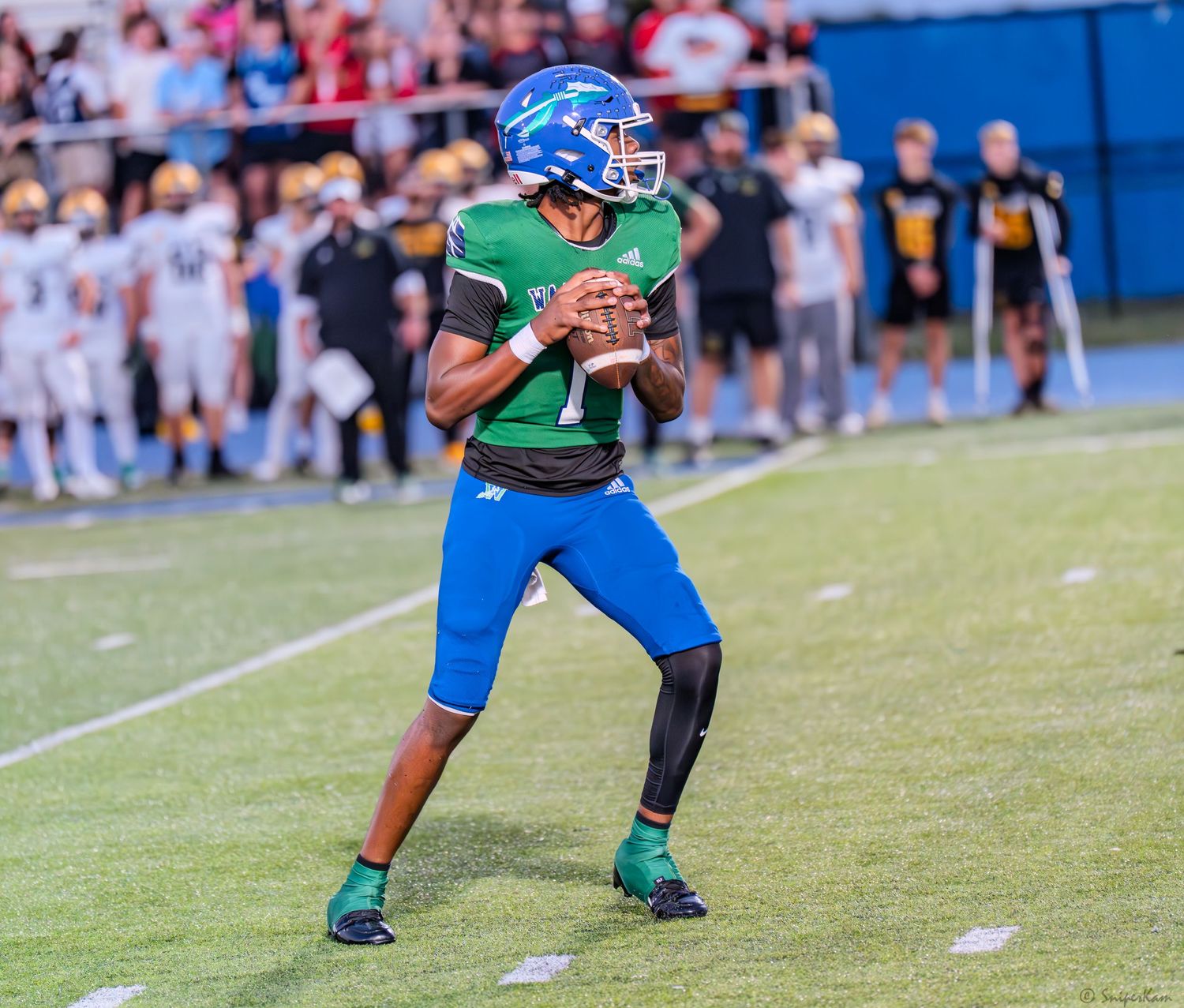 Warriors quarterback number 7 in green and blue uniform prepares to throw during a high school football game.