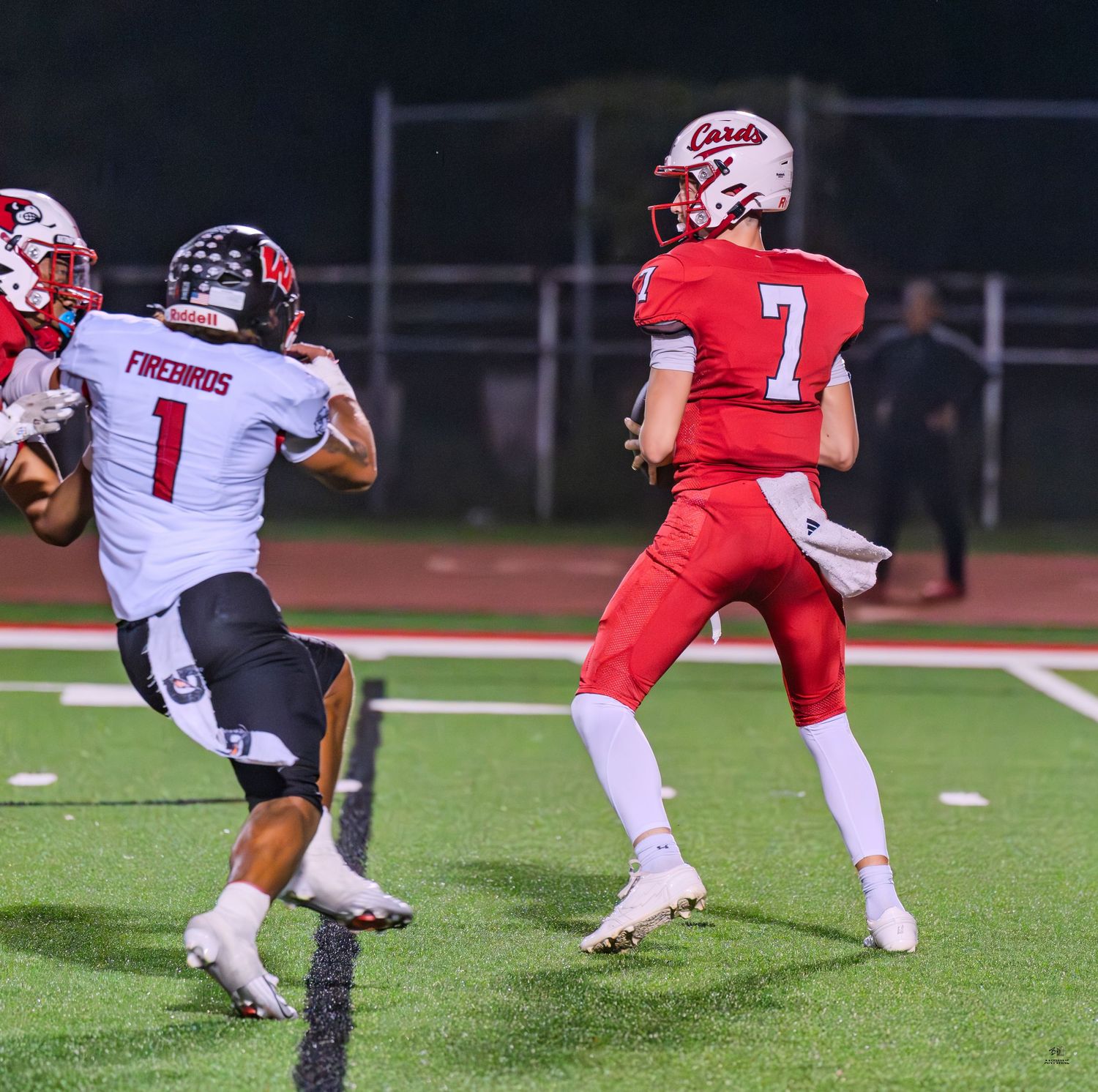 Red-uniformed quarterback number 7 prepares to throw as a Firebirds defender rushes during a night game.